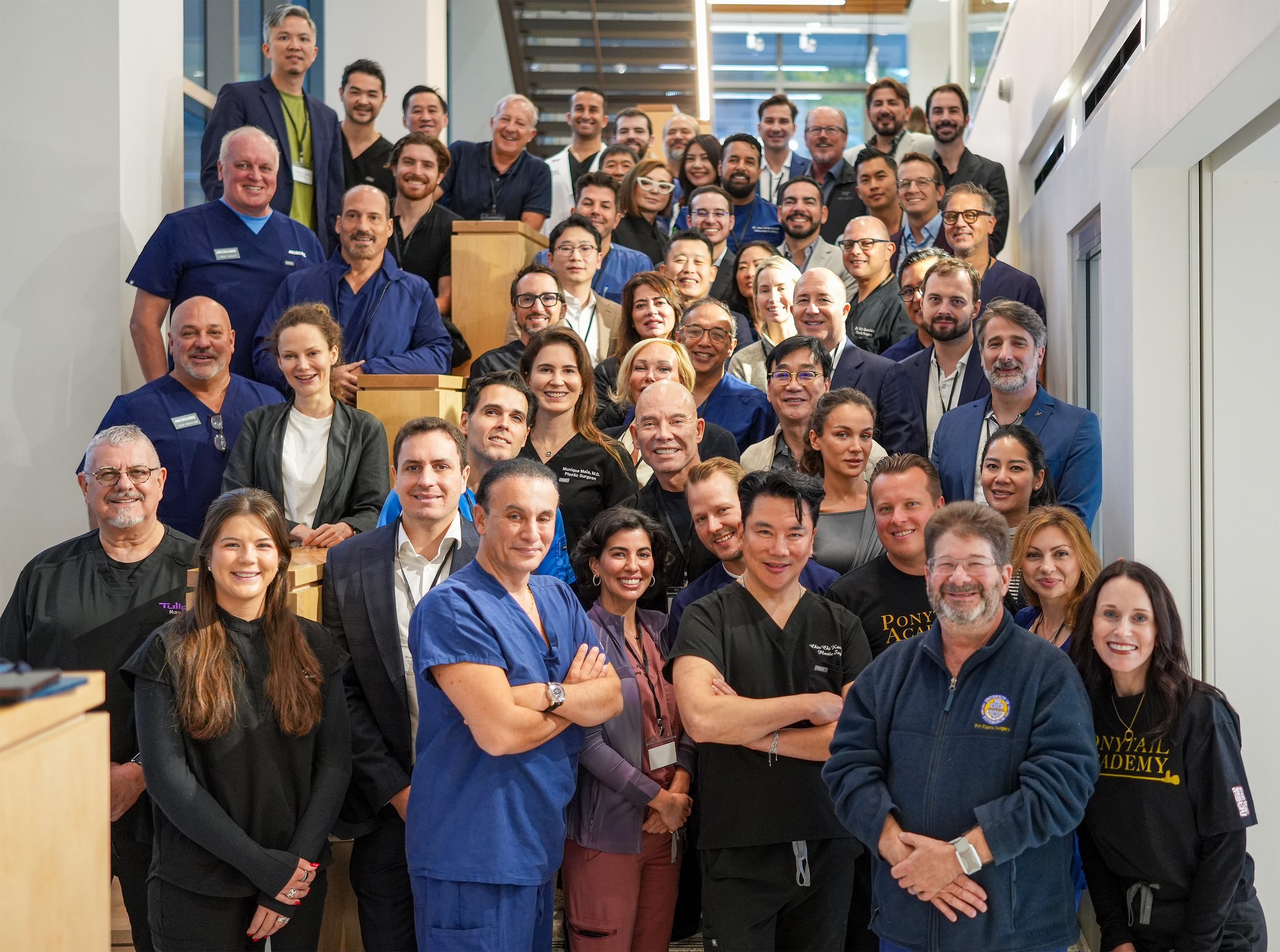 A large group of people gathered on a staircase indoors, smiling for a group photo. The group includes men and women of various ethnicities dressed in professional clothing, some in medical scrubs, some in business attire.