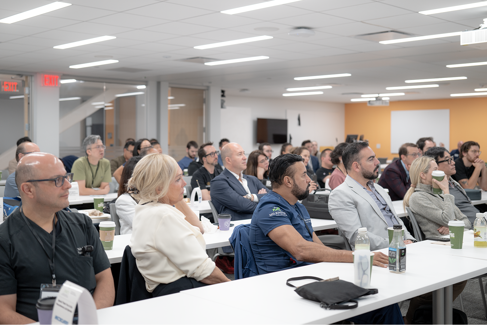 Audience attending a conference in a modern classroom with white tables, bottles and cups, and a yellow accent wall at the back.
