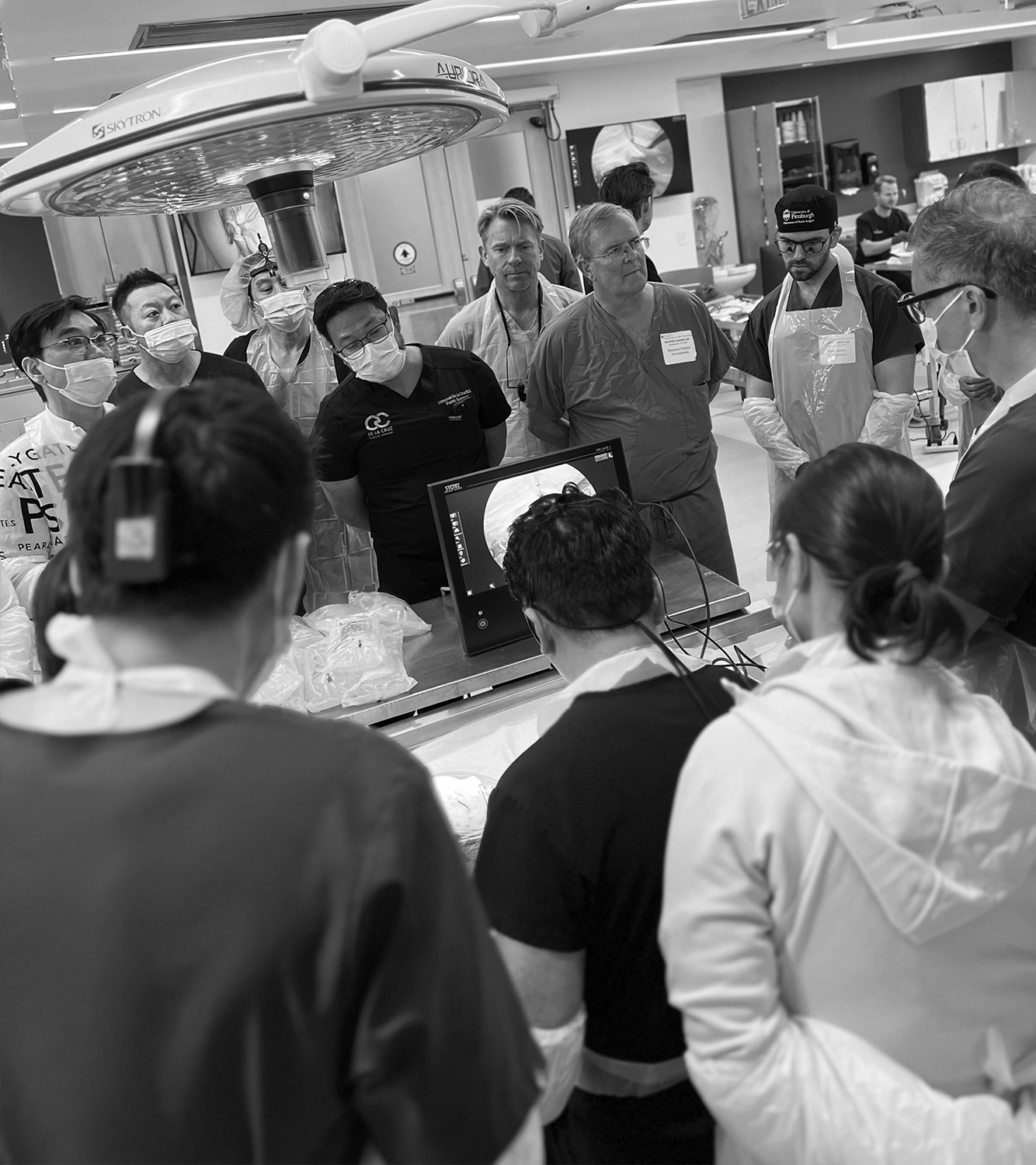 Medical professionals and students gathered around a table in a clinical setting, observing a demonstration on a monitor, all wearing masks and protective gear.