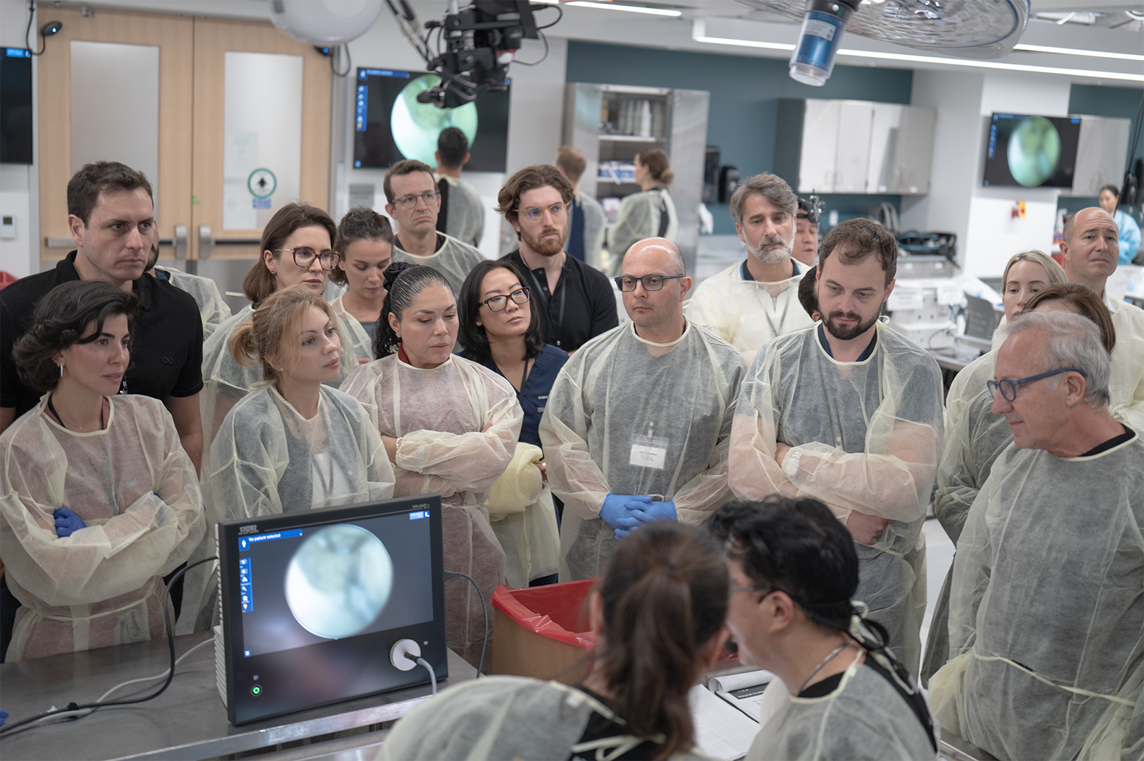 Group of people wearing lab coats and gloves gathered around a table in a medical or scientific laboratory, observing a demonstration or presentation.