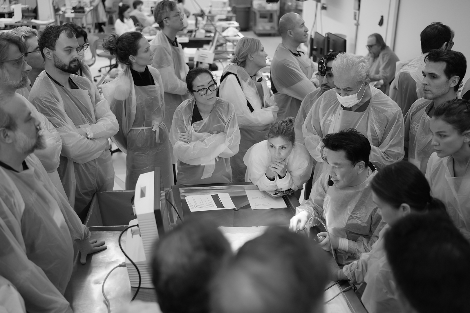 Group of medical professionals and students gathered around a patient in a hospital or clinical setting, with some wearing masks and examination gowns, focused on a procedure.