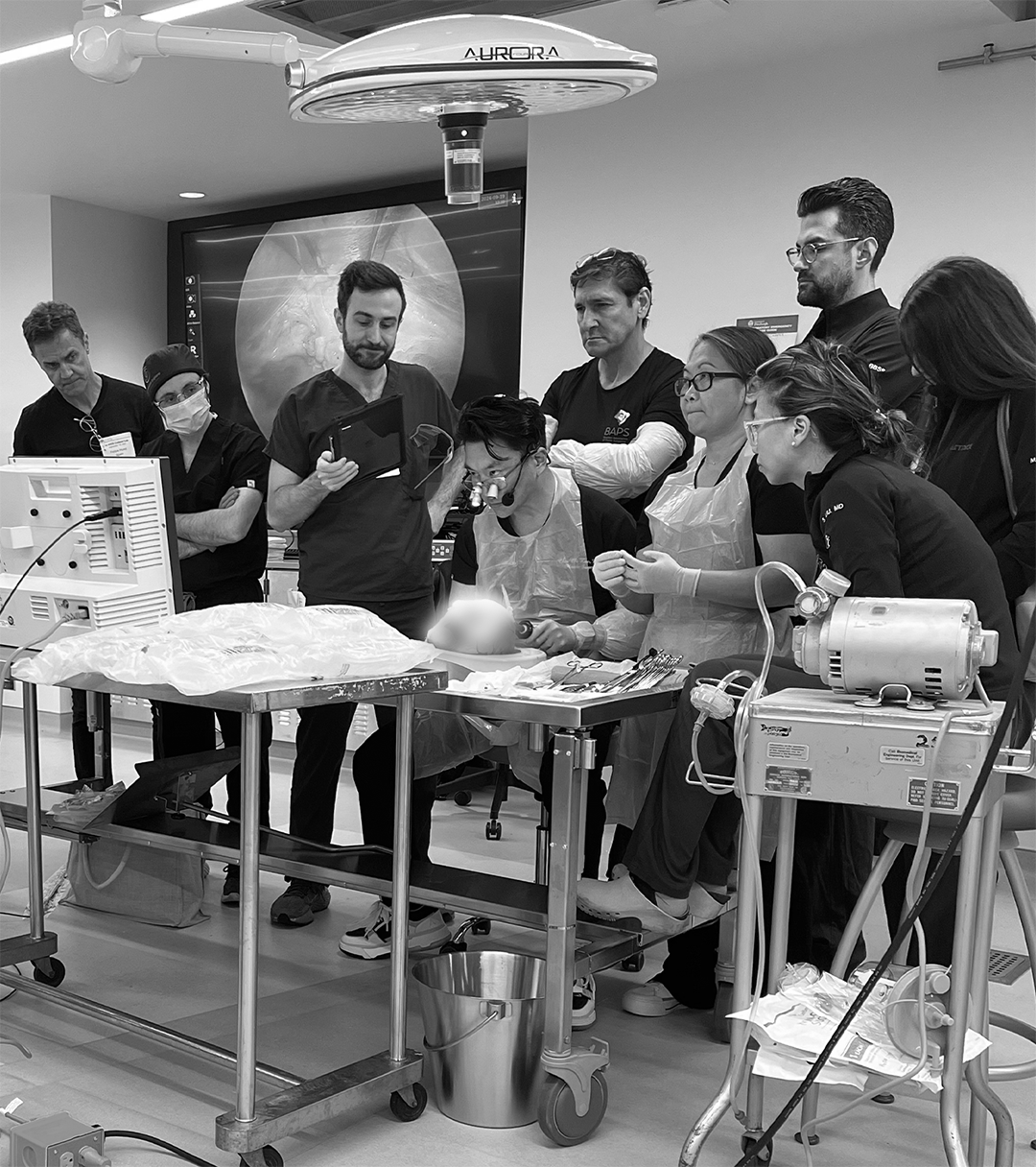 Medical team performing a surgical procedure in an operating room, with doctors and nurses gathered around a patient on the operating table, medical equipment and monitors visible.