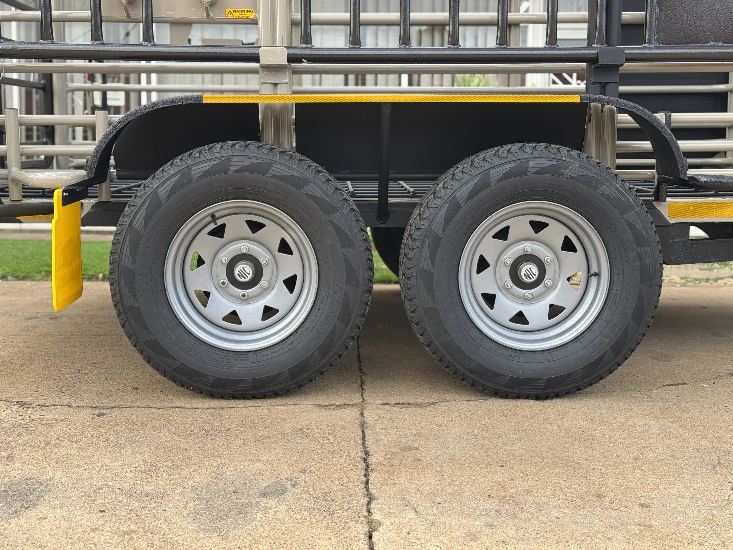 Close-up of two black rubber tires with metal rims on a vehicle trailer. The trailer features a metal frame with yellow reflective strips and a small yellow mudguard. The background includes paved ground and a grassy area.