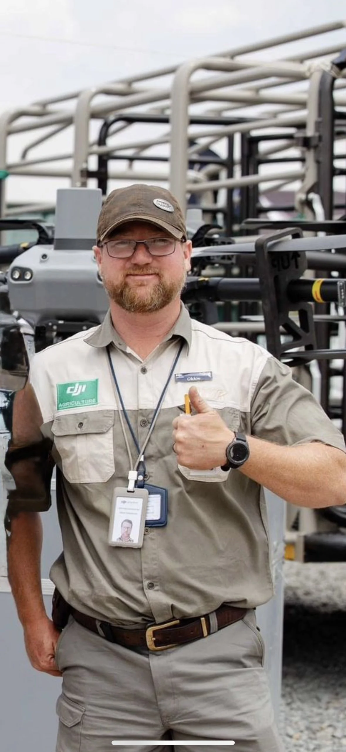 Man in beige work uniform, wearing a brown cap and glasses, giving a thumbs-up, standing in front of equipment with metal frames, likely at a work site.