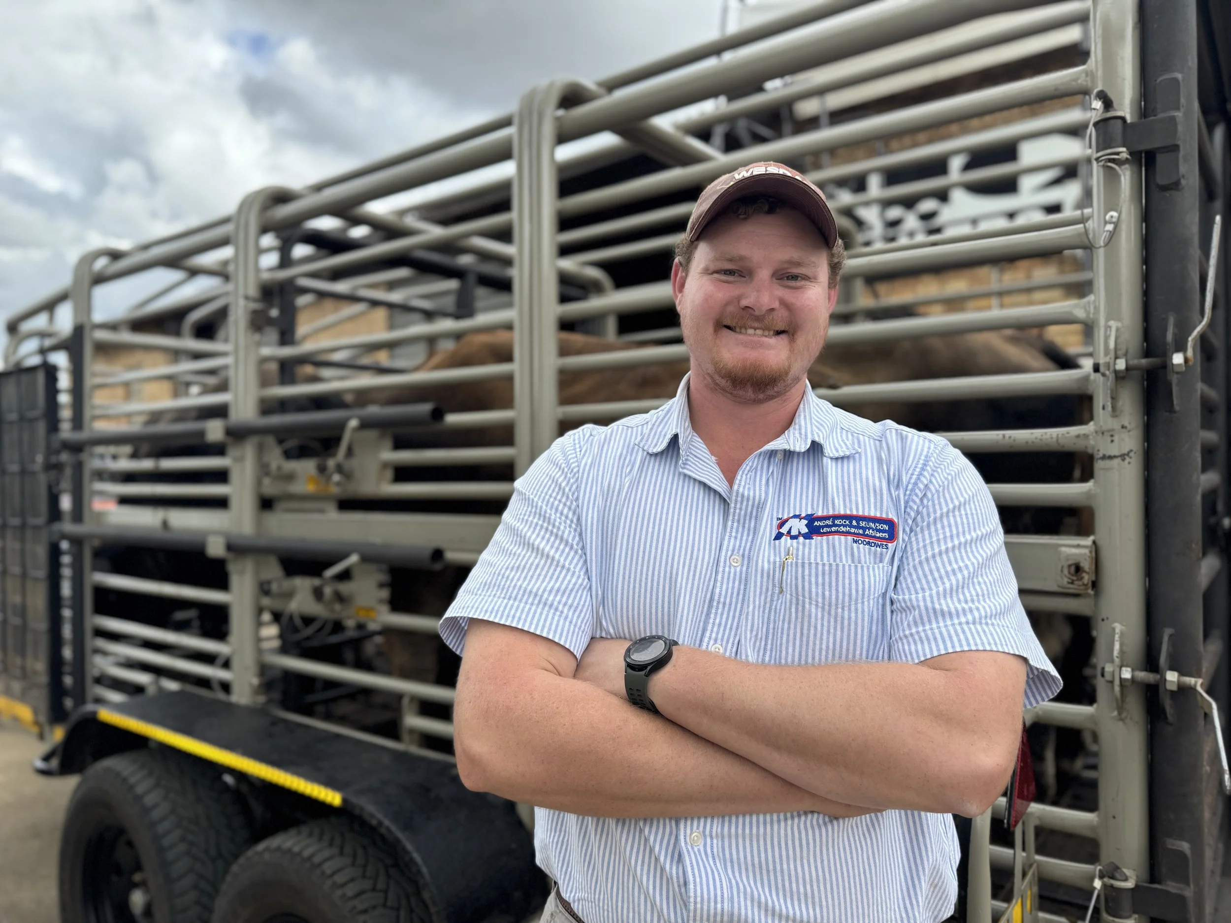 A man standing with arms crossed in front of a livestock trailer, smiling, with a brown cow visible inside the trailer.