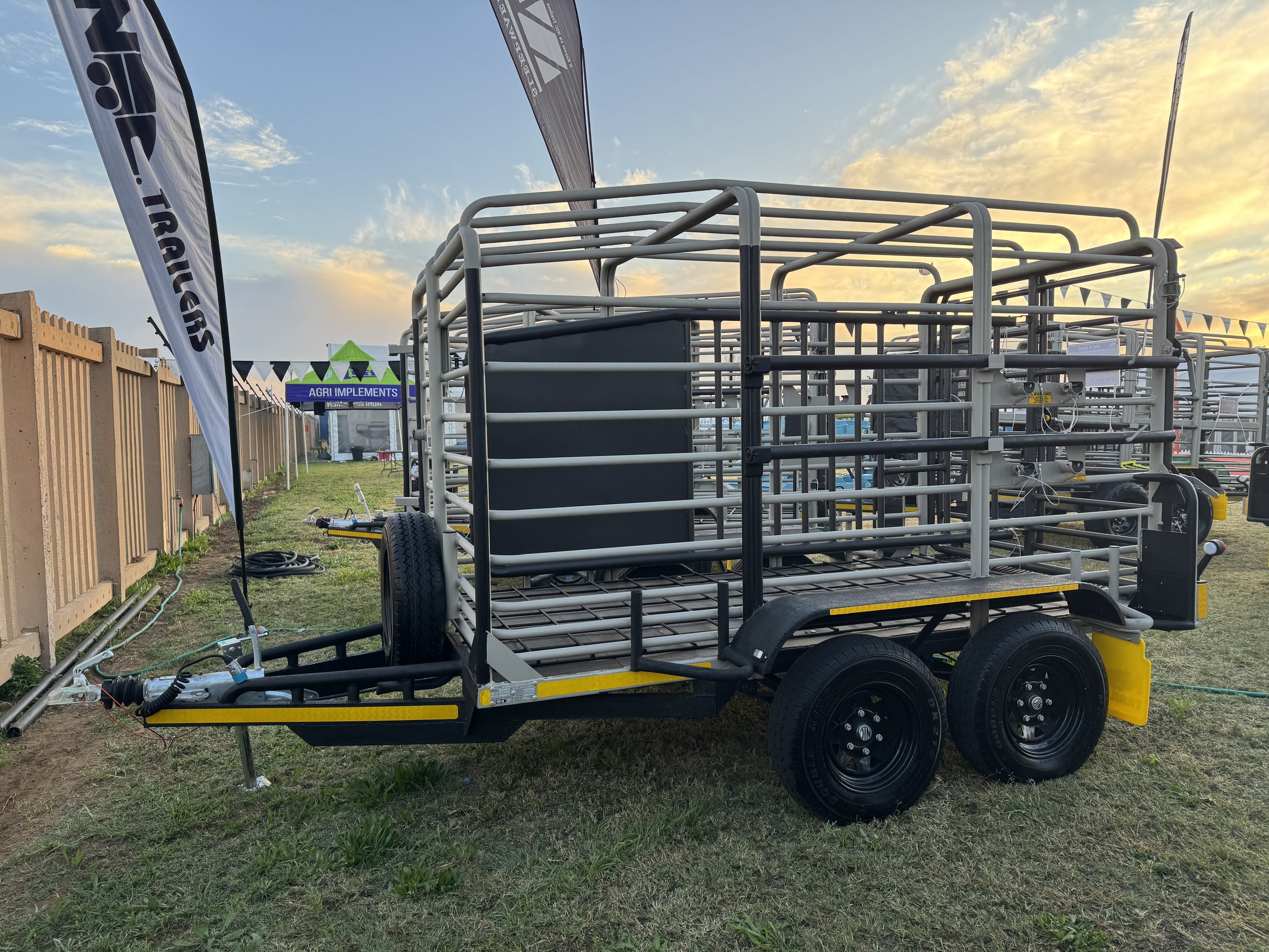 Empty livestock trailer parked on grass at outdoor event, with flags and tents in the background during sunset.