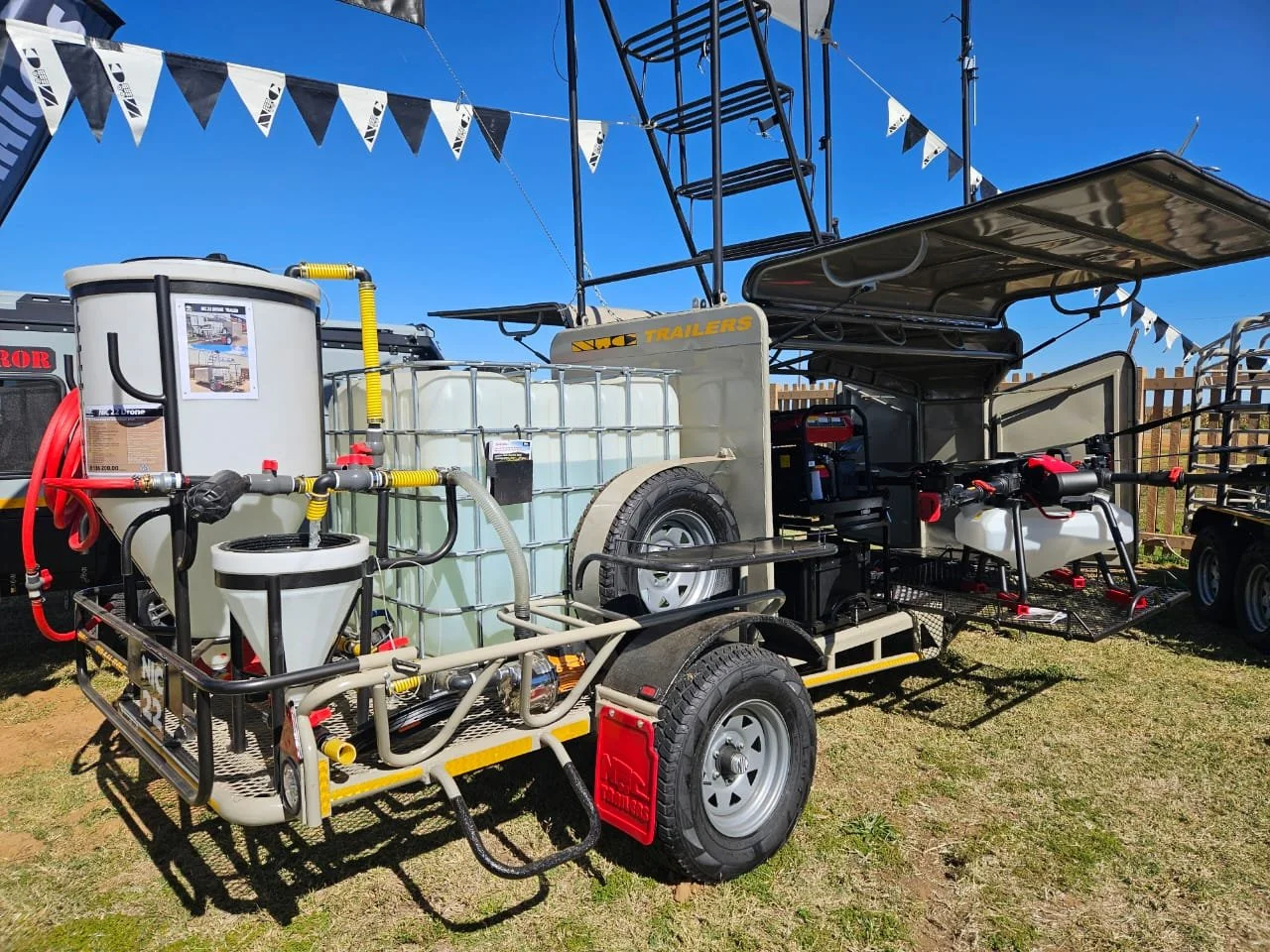 A portable fire fighting water trailer equipped with tanks, hoses, spray nozzles, and spraying equipment, set on a grassy field under a clear blue sky.