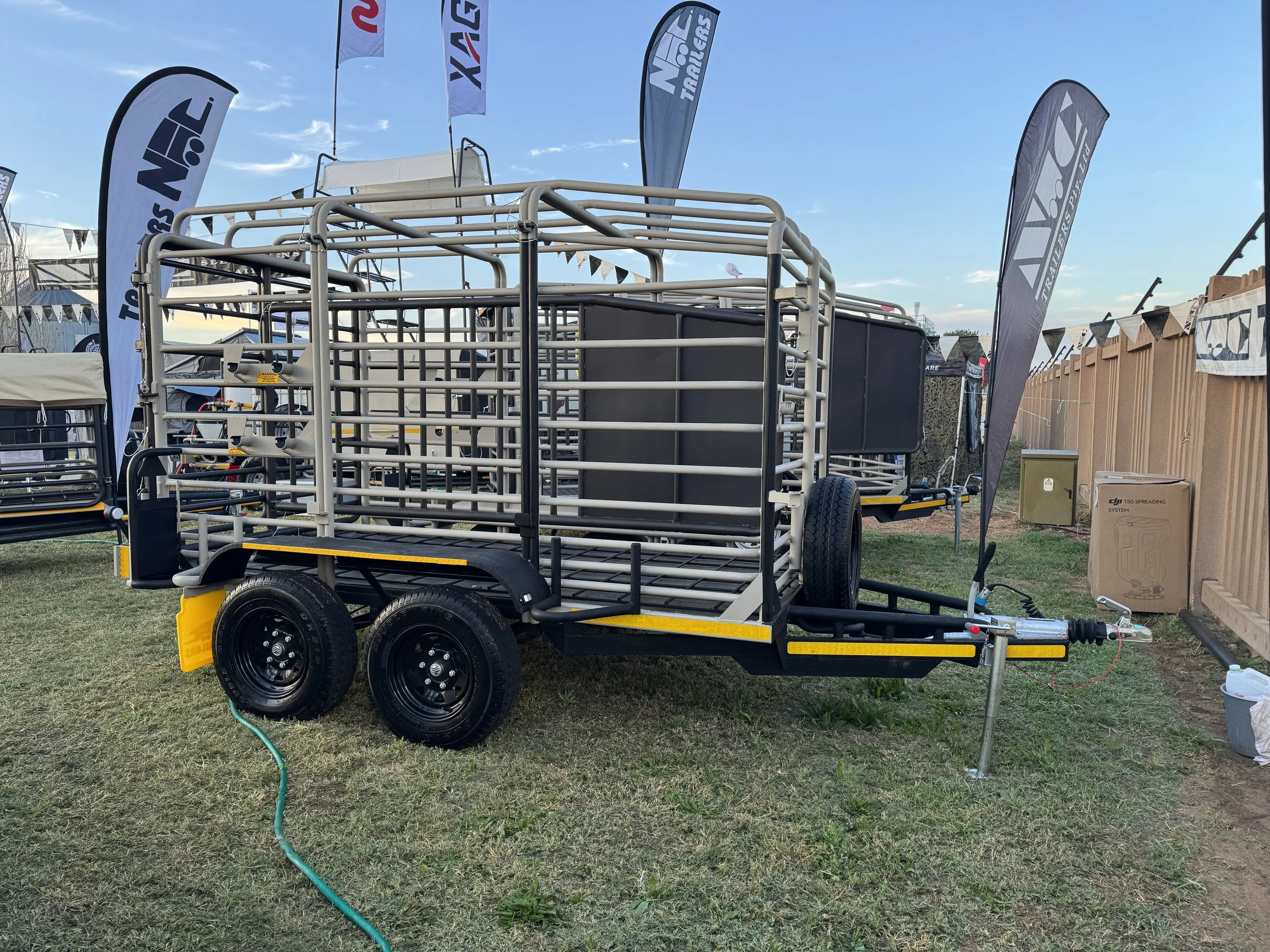 A livestock trailer with double axles and metal fencing, parked on grass at an outdoor event with flags and banners in the background.