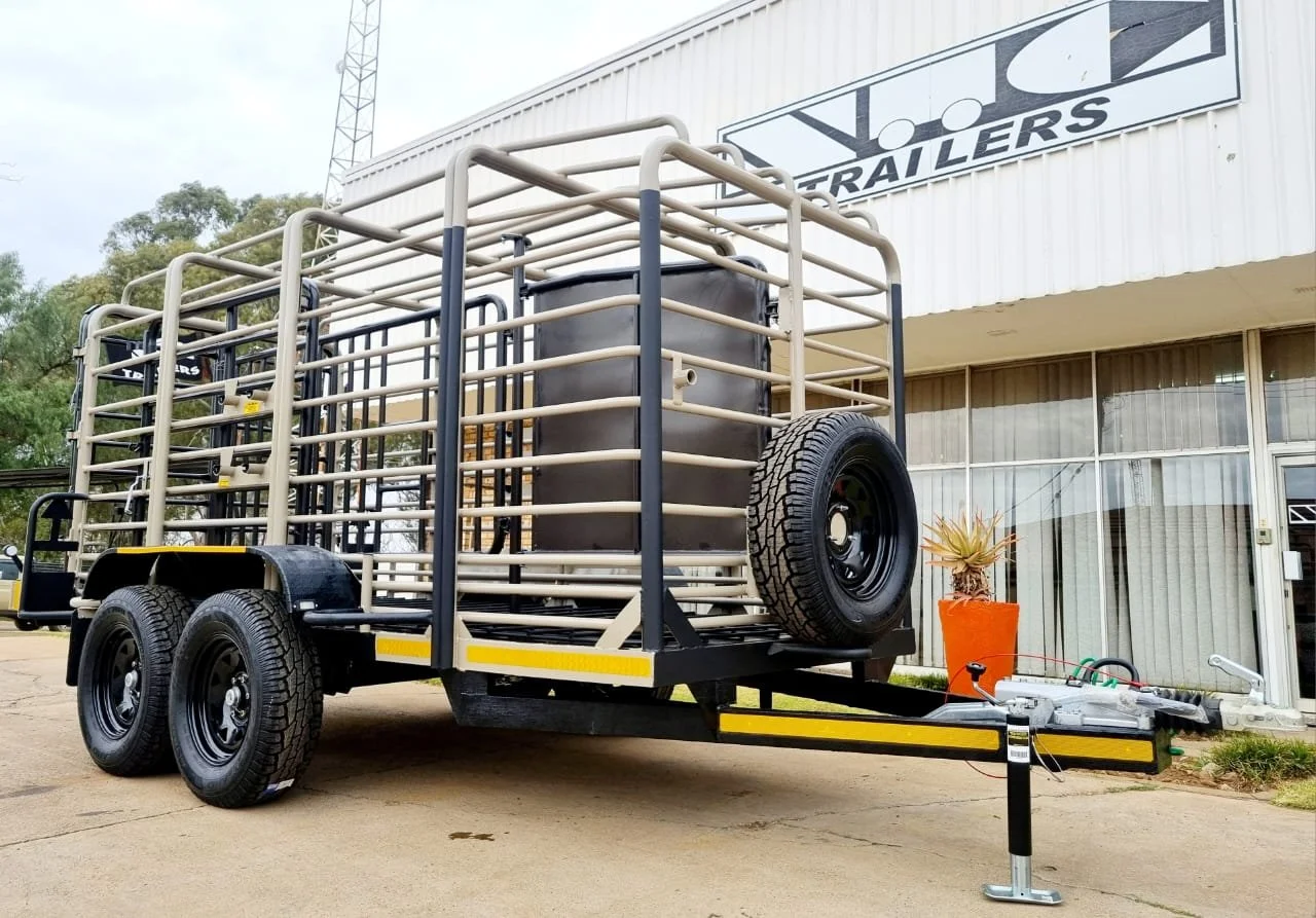 Heavy-duty cattle and sheep trailer with metal framework and spare tire, parked outside a building with signage. Double axle construction and a tow hitch are visible.