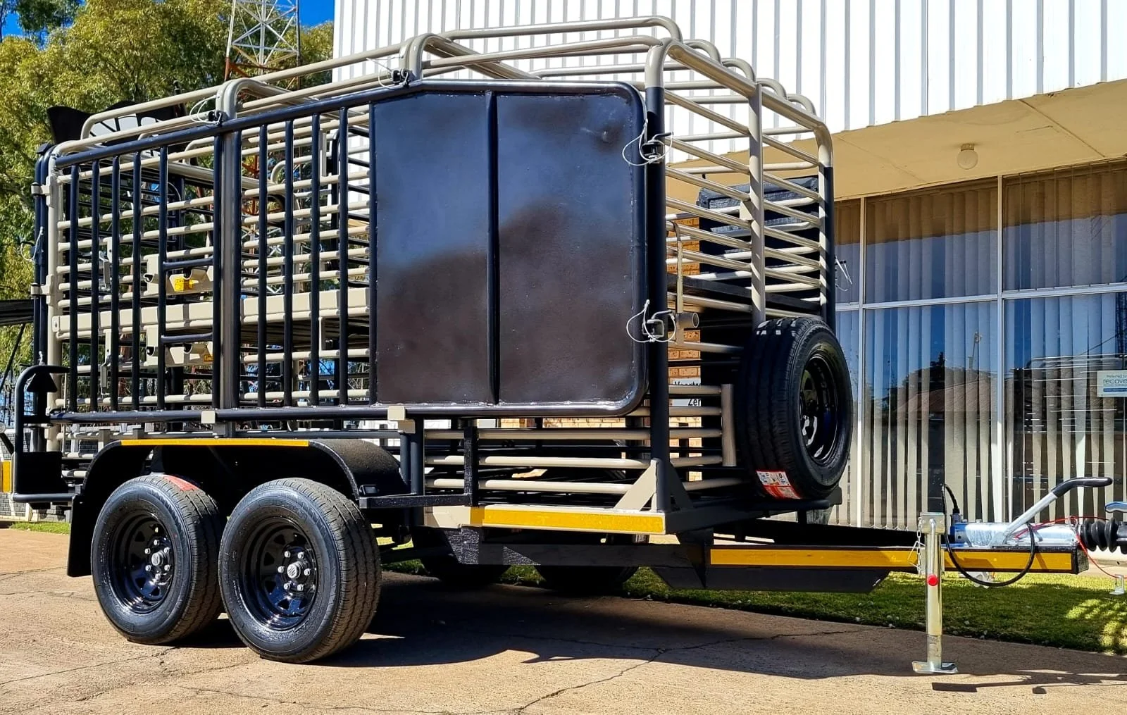 A black utility trailer with dual wheels, containing metal cages and racks, parked outside near a building with large glass windows and a grassy area.