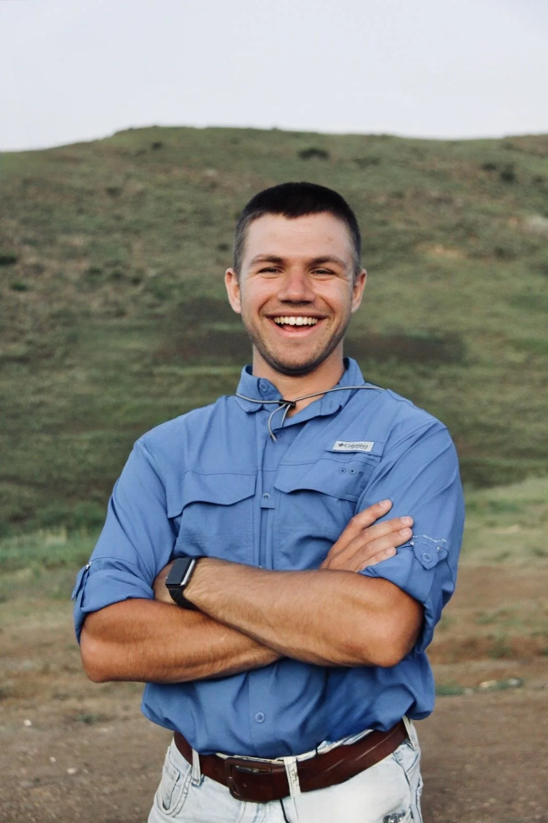 A smiling young man with crossed arms standing outdoors in front of a grassy hillside, wearing a blue Columbia shirt and a smartwatch.