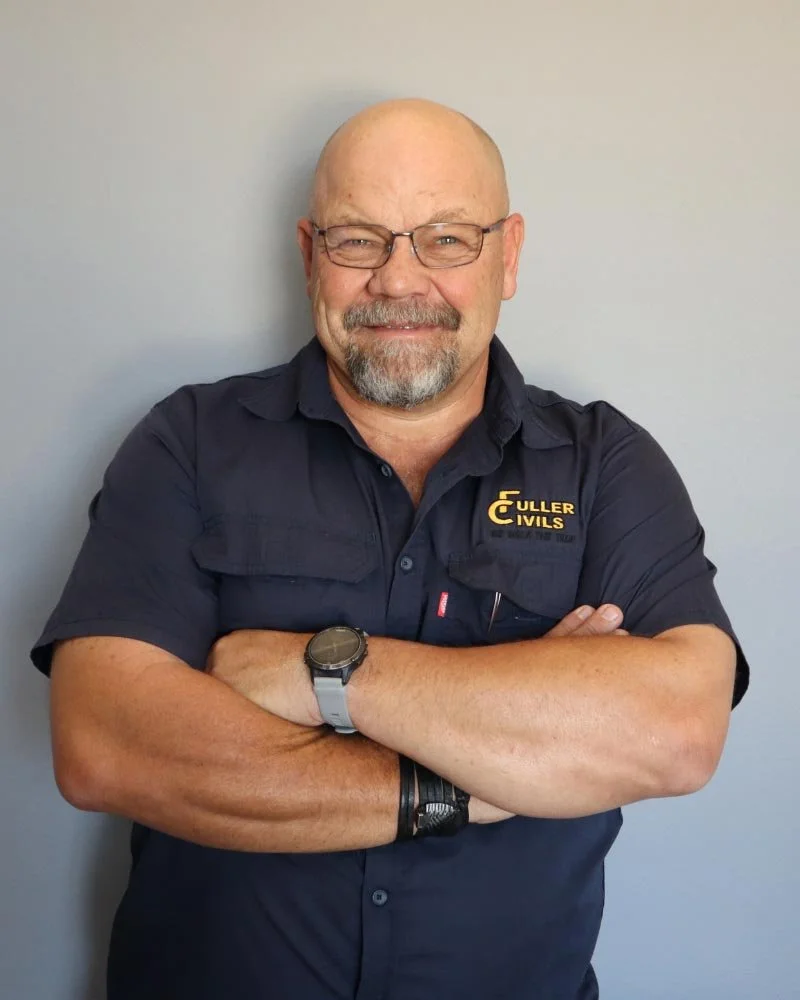 A smiling man with a bald head, grey beard, and glasses standing with arms crossed in front of a plain grey wall. He is wearing a black short-sleeved shirt with a logo that reads 'Fuller Civils' in yellow and white text.