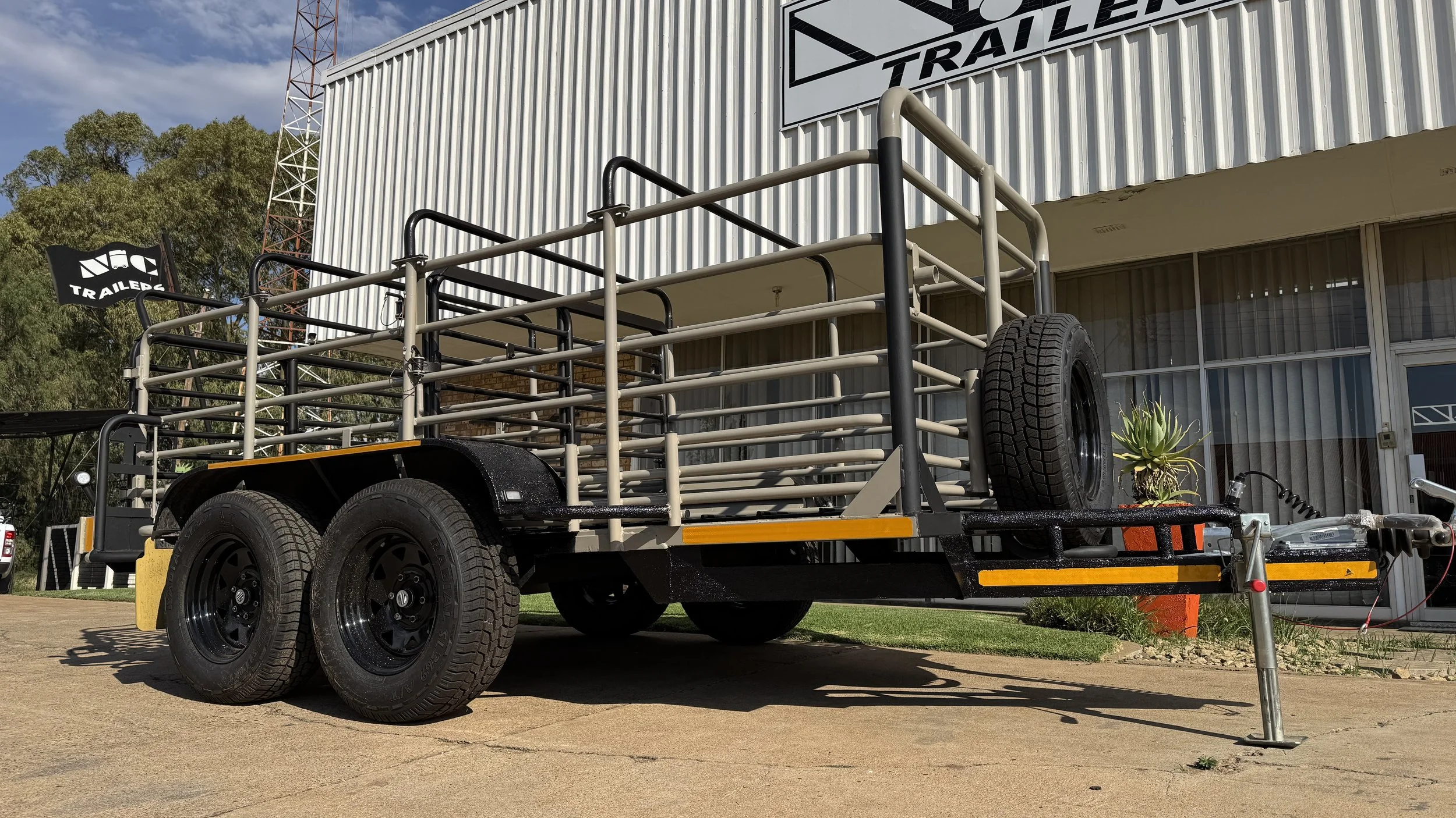 Livestock trailer with a dual-axle design parked outside a building with signage, featuring metal railings and a mounted spare tire.