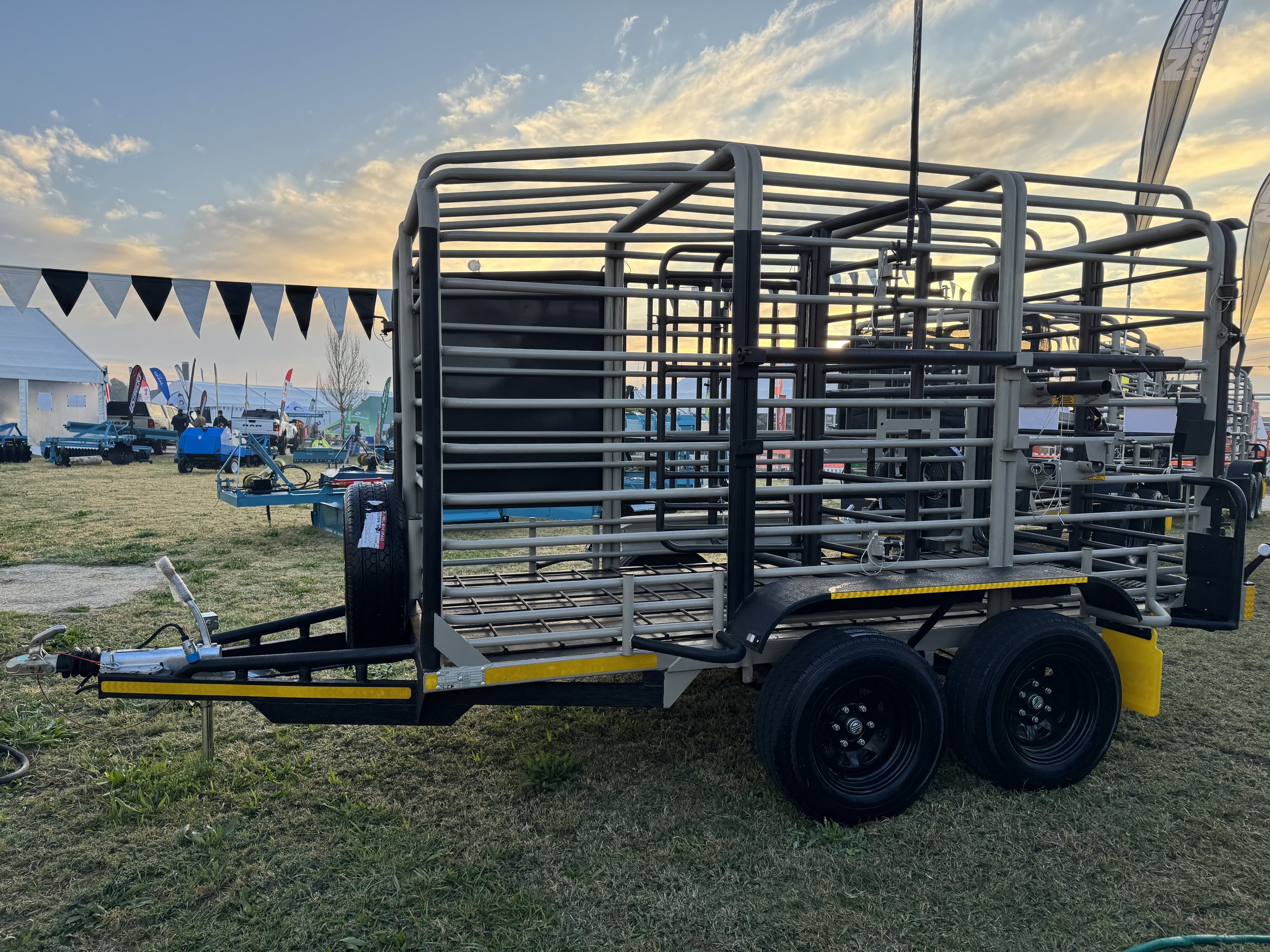 A livestock trailer parked on grass under a sunset sky, with other trailers and tents visible in the background.