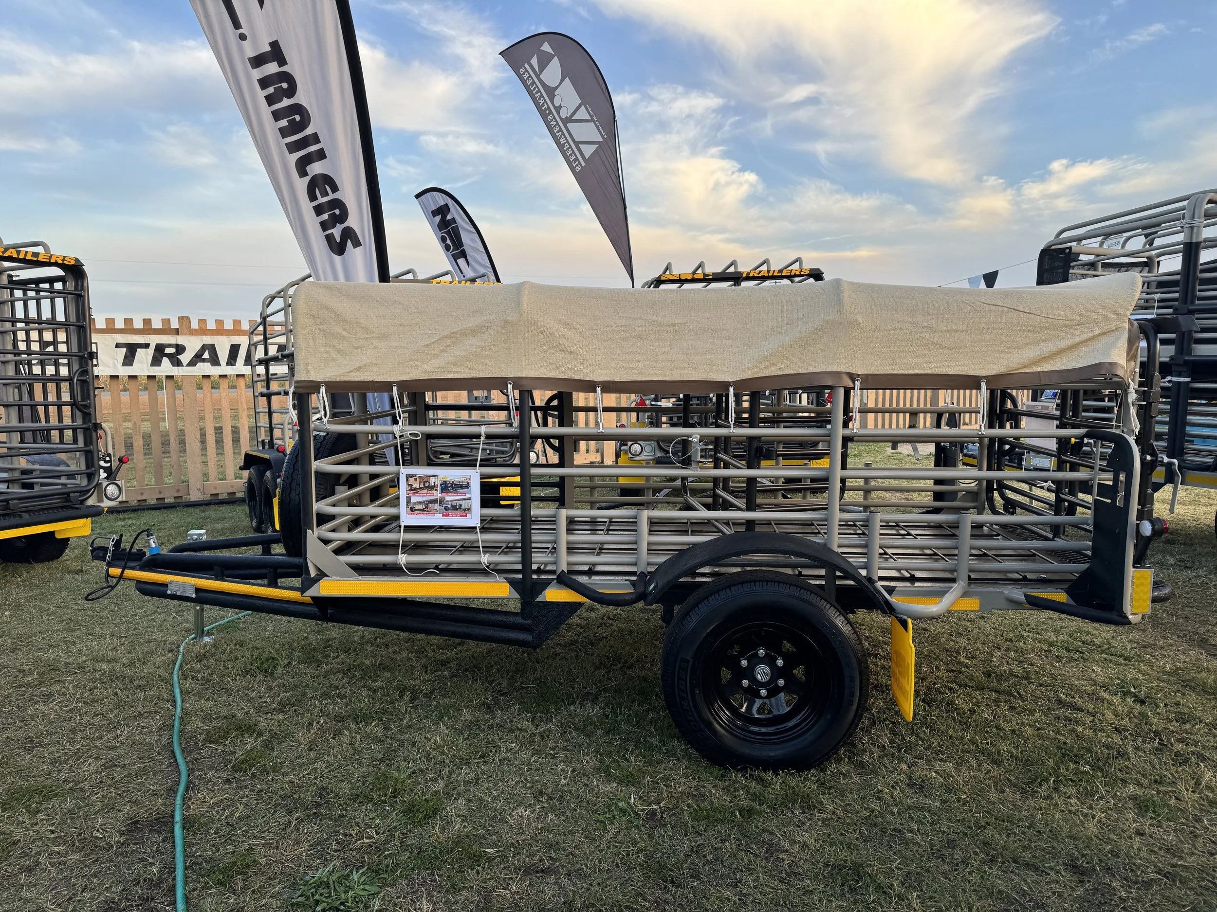 A metal livestock trailer with a beige cover, parked outdoors on grass during sunset, surrounded by flags and other trailers.