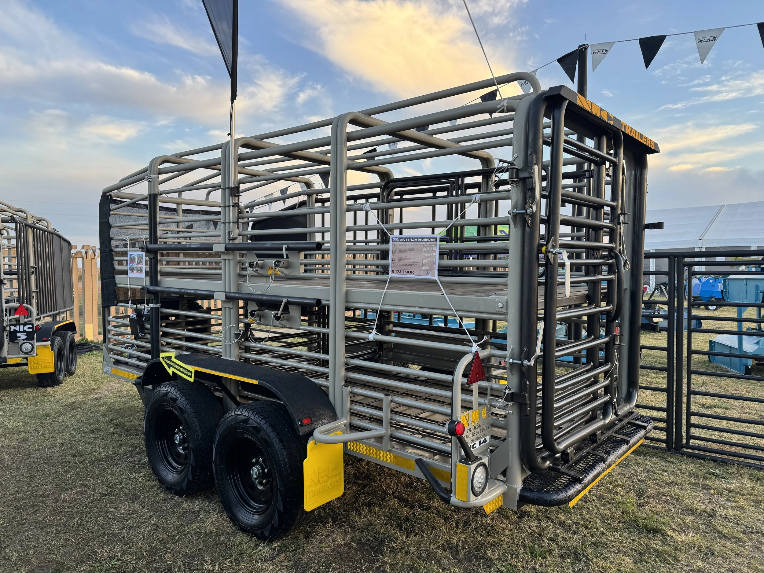 Large metallic livestock trailer with dual axles on grass field at sunset, surrounded by other trailers and fencing