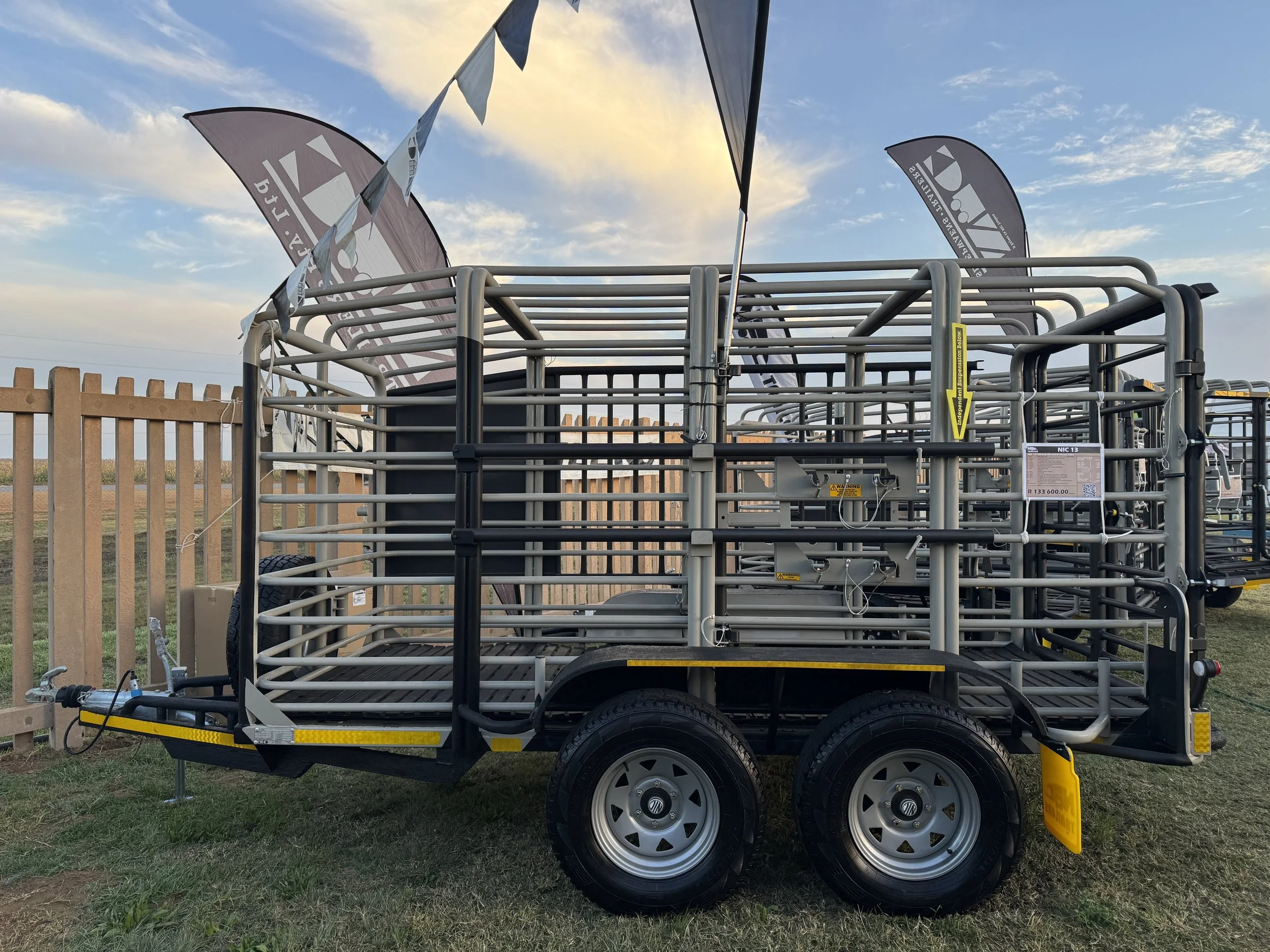 A large metal livestock trailer with dual wheels parked on grass near a wooden fence and flags waving in the distance, under a partly cloudy sky during sunset.