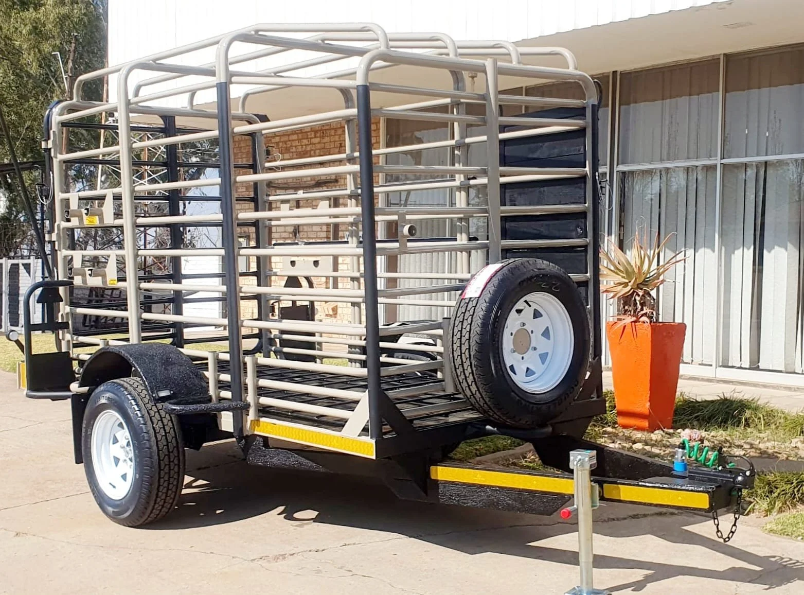 A metal livestock trailer with white wheels parked outside a building with large windows.