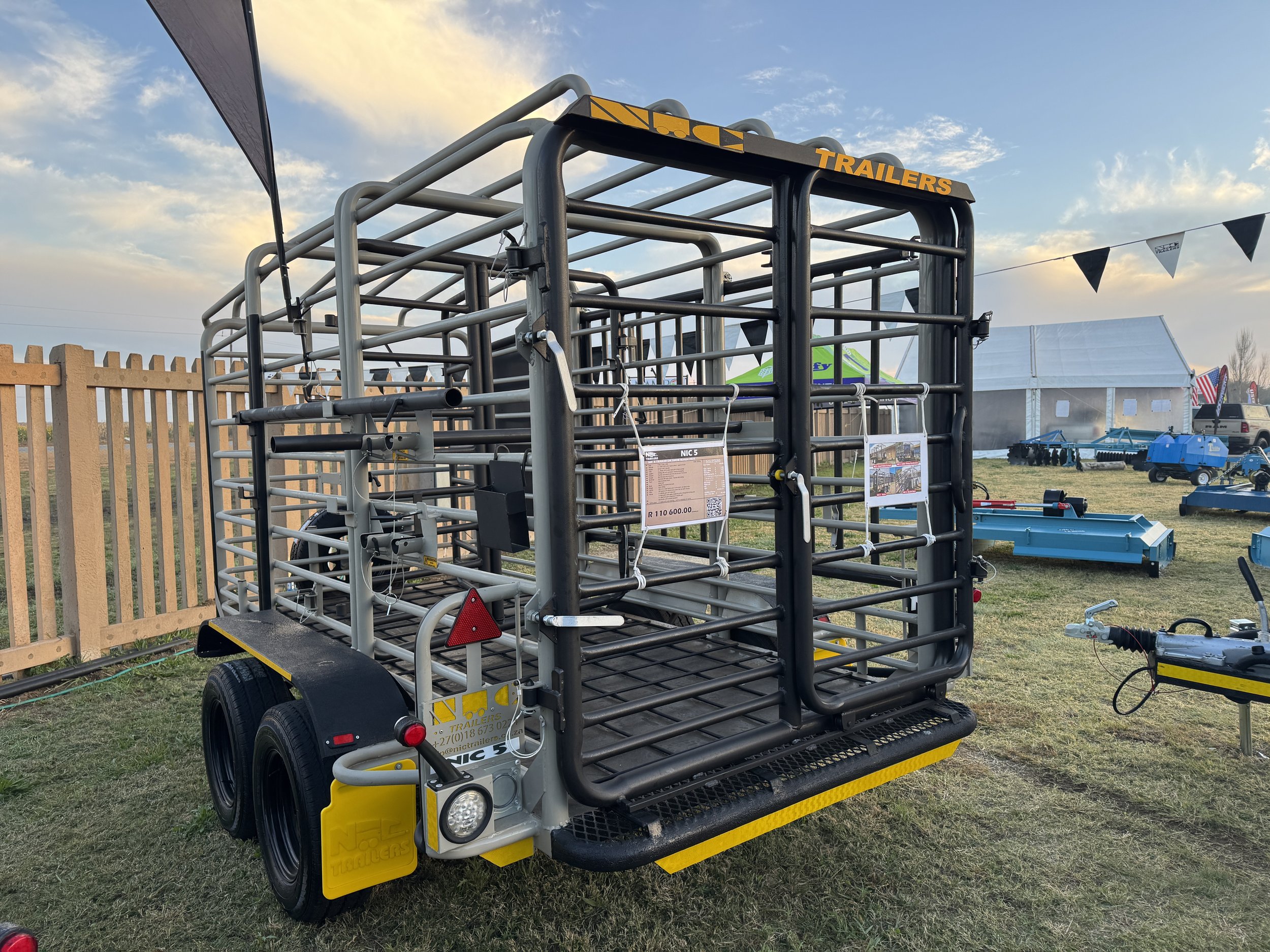 A livestock trailer with metal railings parked on grass at an outdoor event, with tents and other equipment visible in the background during sunset.
