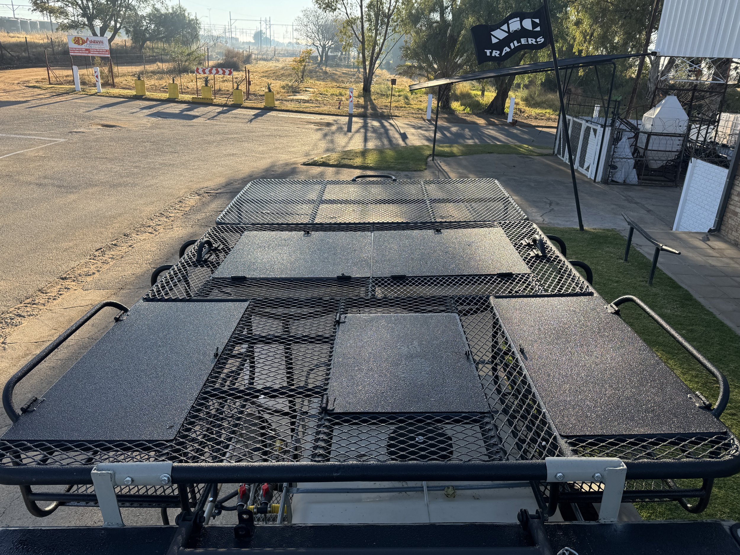A flatbed trailer with a metal mesh surface and black panels, parked outdoors near a sidewalk and grassy area, with trees and a sign in the background.