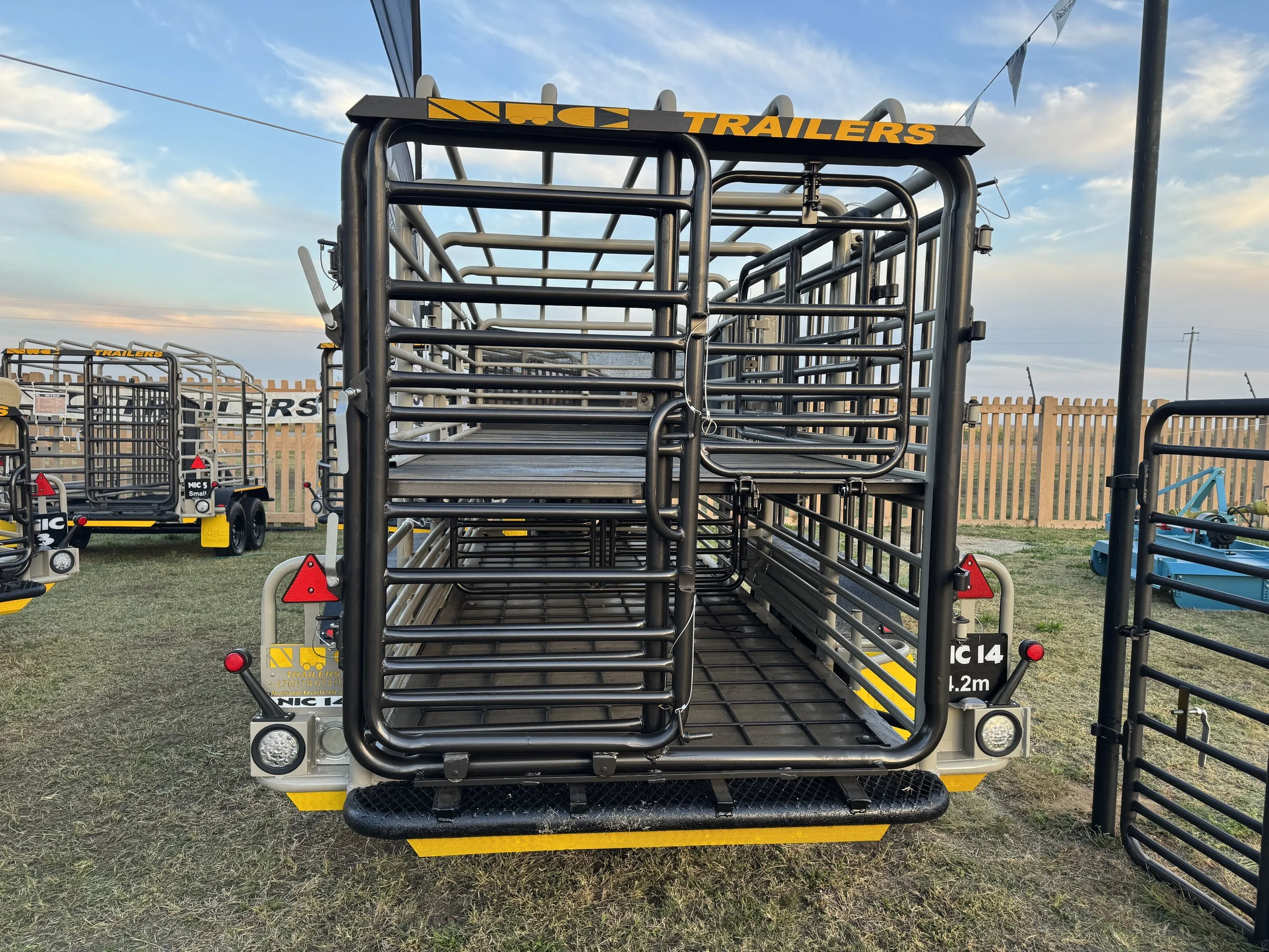A cattle trailer with black metal bars, parked on grass with other trailers nearby, under a partly cloudy sky.