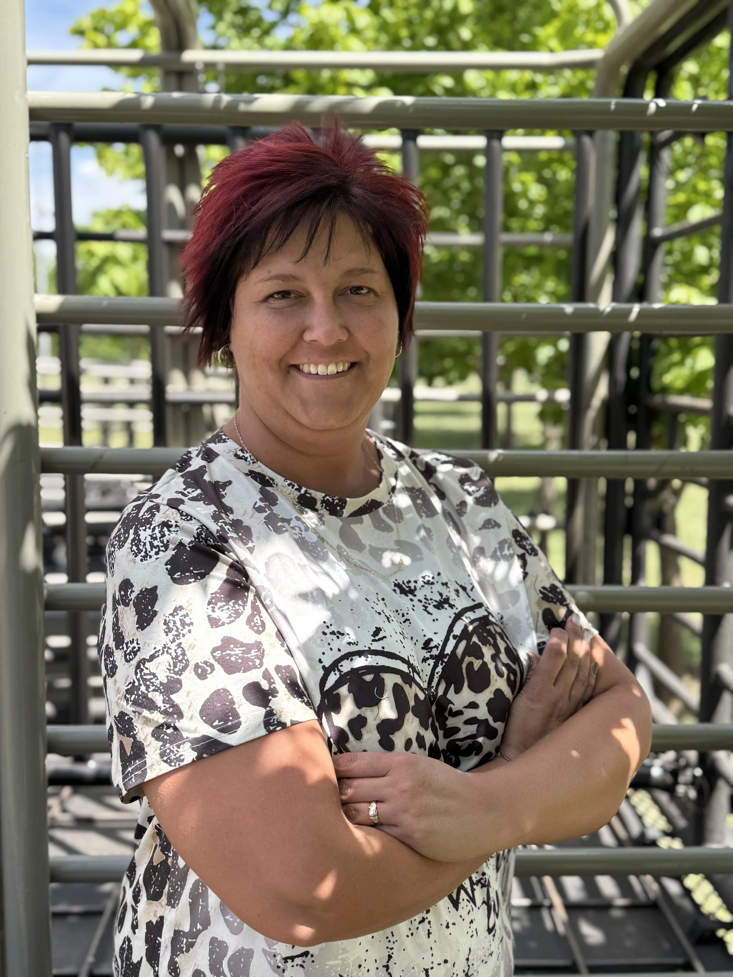 A woman with red and black hair smiling with arms crossed, standing outdoors in front of a metal playground structure with trees in the background.