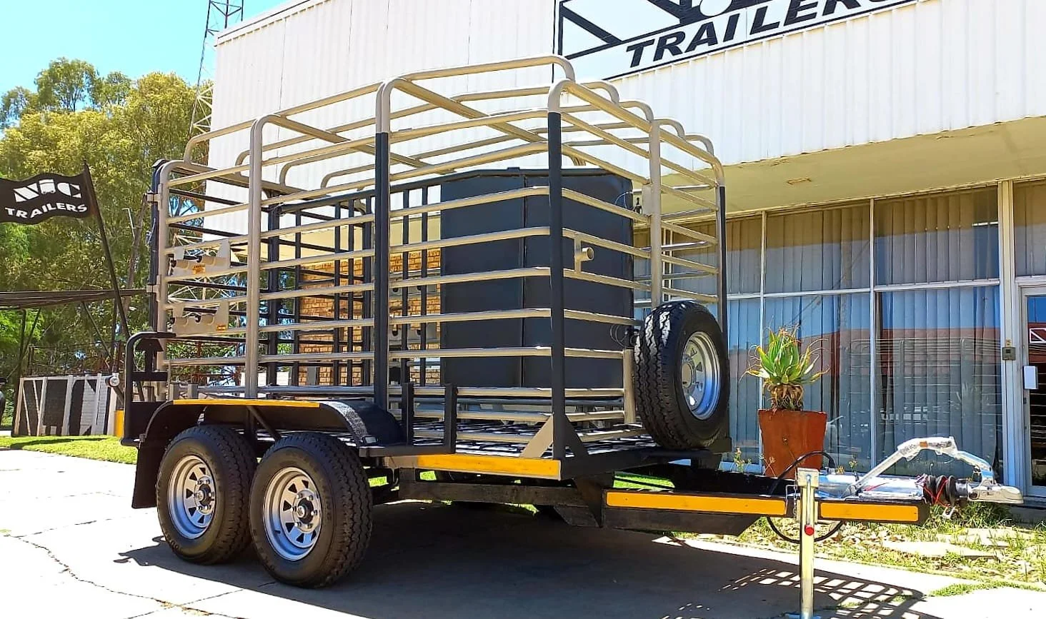 A trailer with a metal frame and two axles parked outside a building with a sign that reads 'Trailers'. The trailer is empty except for a black box and a small potted plant. The background includes a tree and a blue sky.