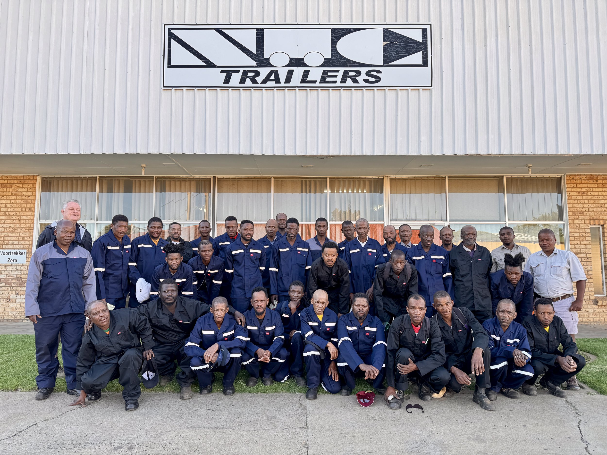 Group of people in uniform posing in front of a trailer company building with a sign that reads 'N.O.C. Trailers'.