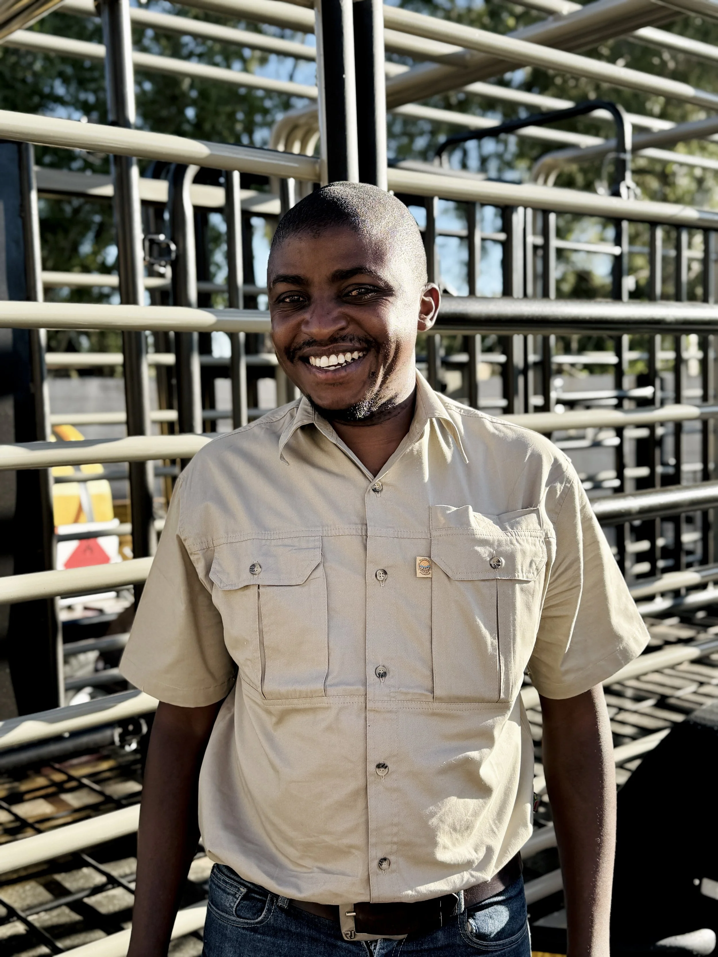 A smiling man in a beige short-sleeve button-up shirt standing outdoors in front of metal railings, with trees and blue sky in the background.