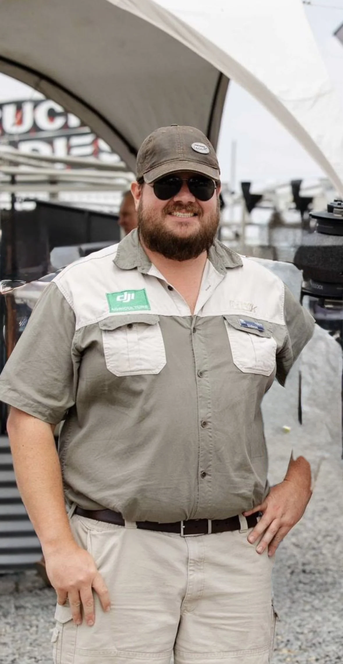A man with a beard and sunglasses standing outdoors, wearing a beige shirt with patches, a cap, and beige shorts, smiling at the camera.