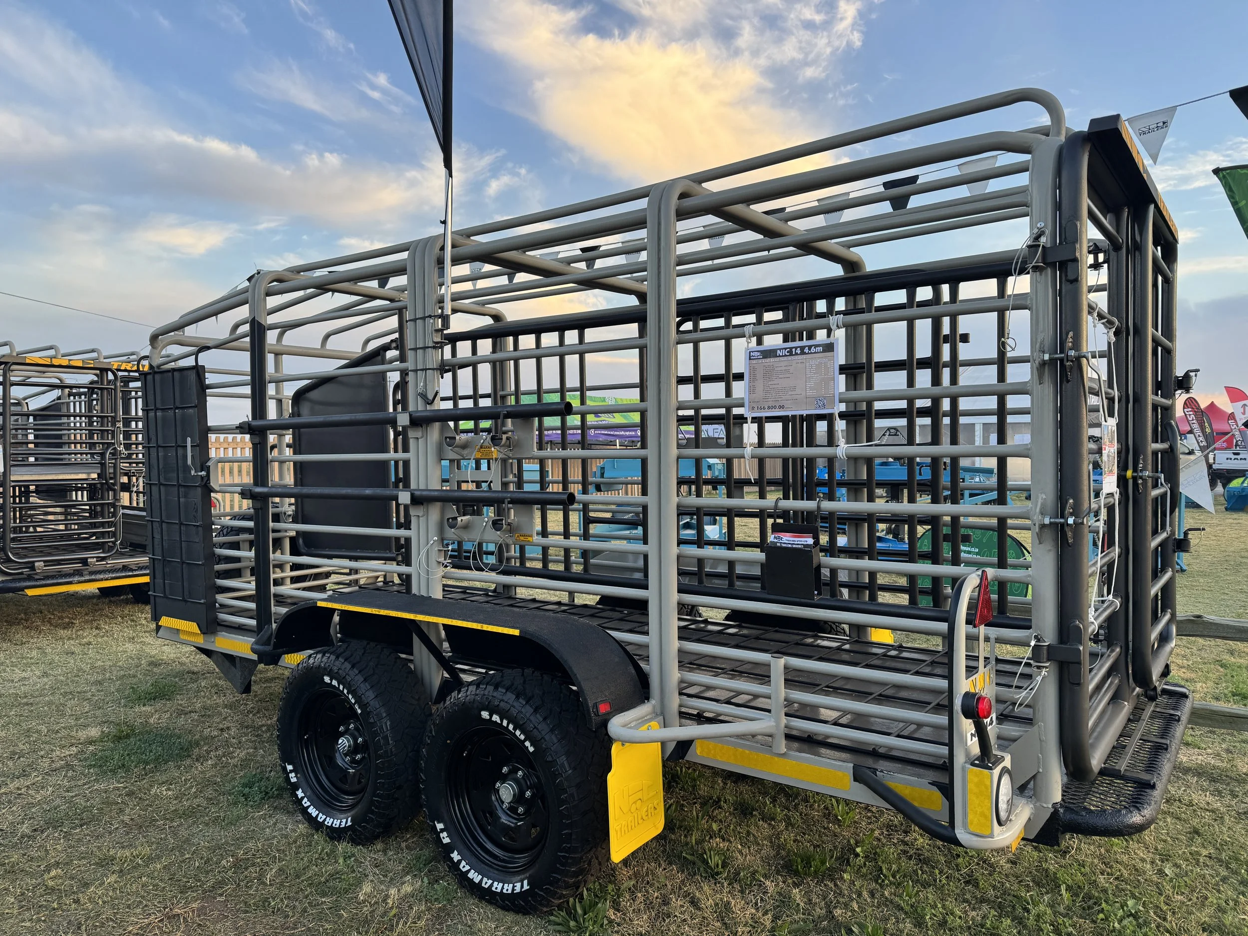 A metal livestock trailer with double axle tires parked on grassy ground at an outdoor event, with other trailers and flags in the background during sunset.
