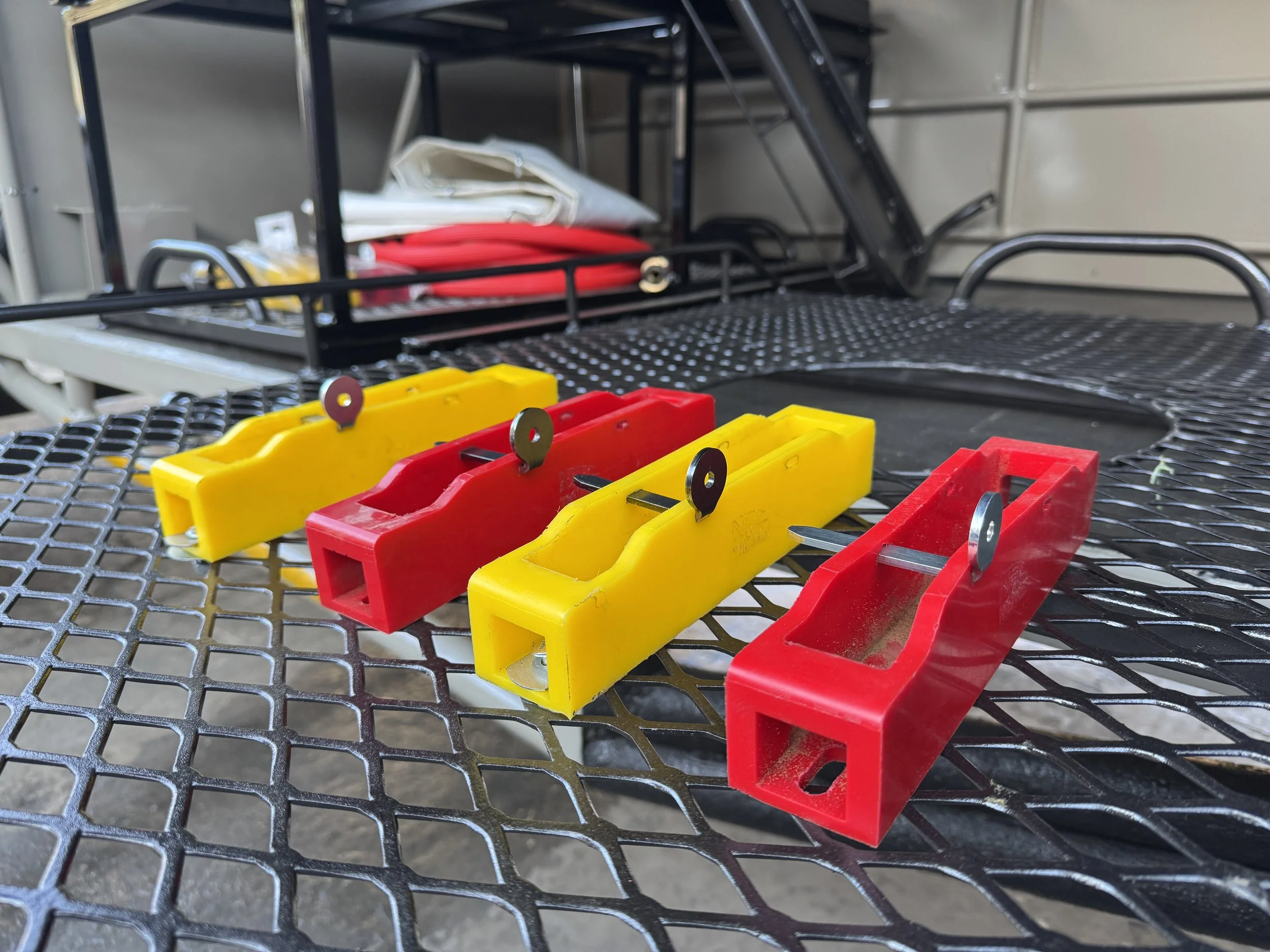 Four red and yellow plastic objects with metal pins on a metal mesh surface, with a cart and various items in the background.