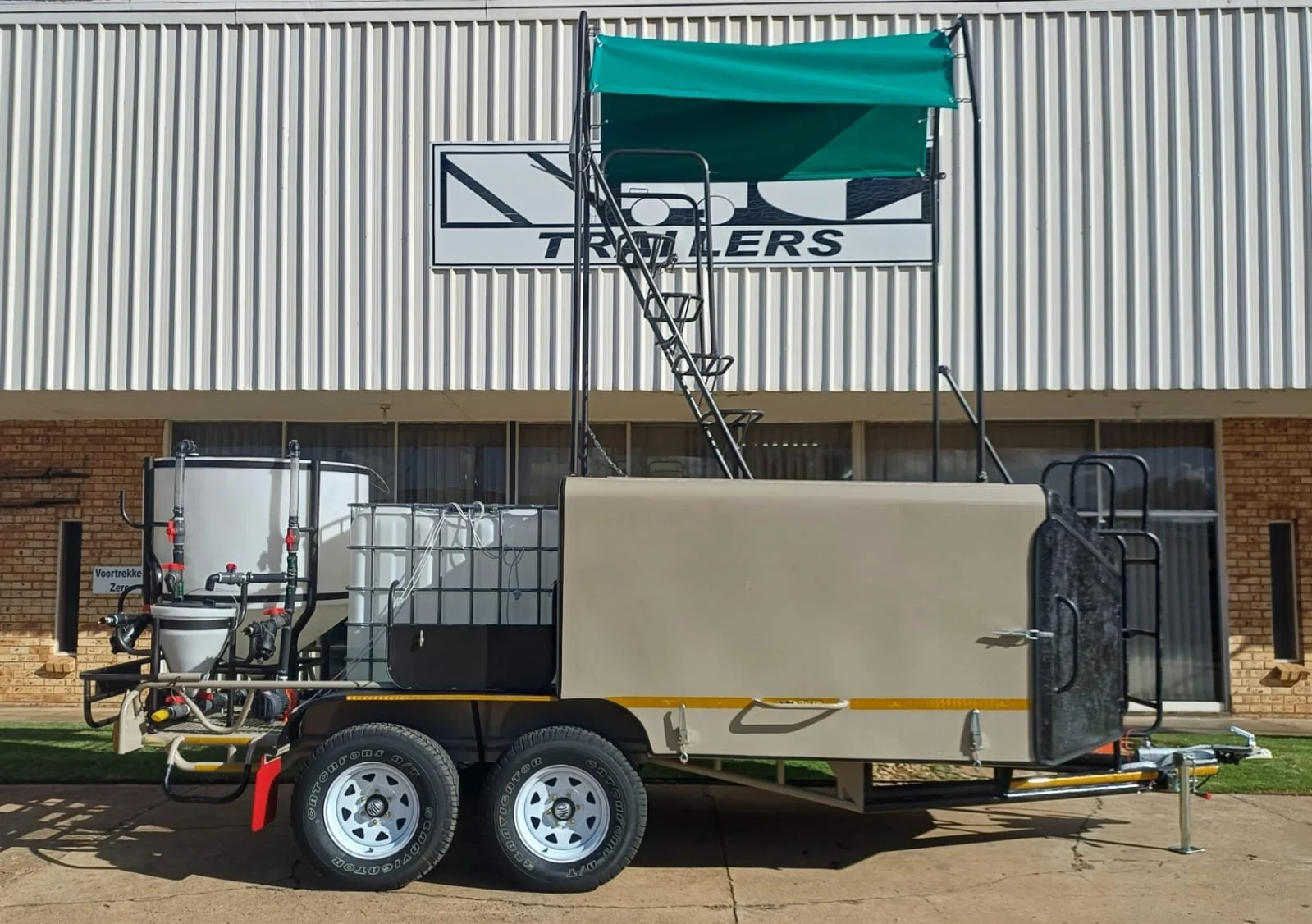 A mobile water treatment and firefighting trailer parked in front of a building with a sign that reads "TRAILERS". The trailer has tanks, hoses, pumps, and a green canopy on a metal frame, with black and white wheels.