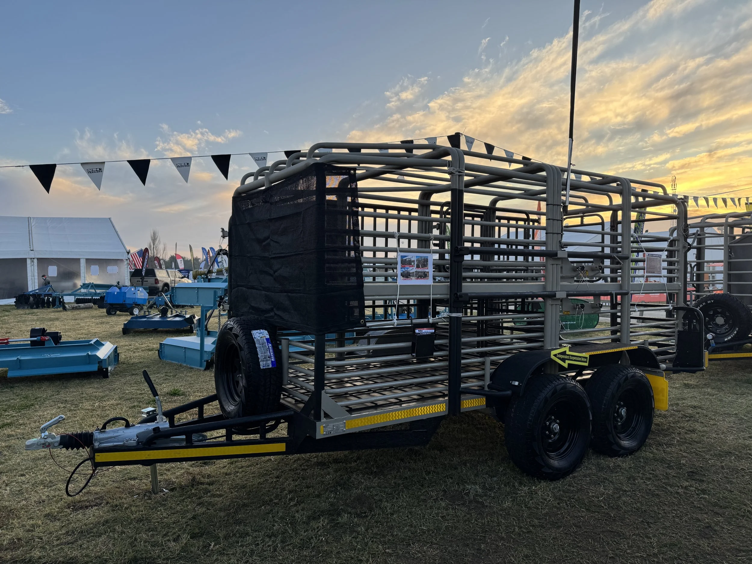 A livestock trailer with metal bars and black tires parked on grass at a fairground during sunset. Other equipment and flags are visible in the background.