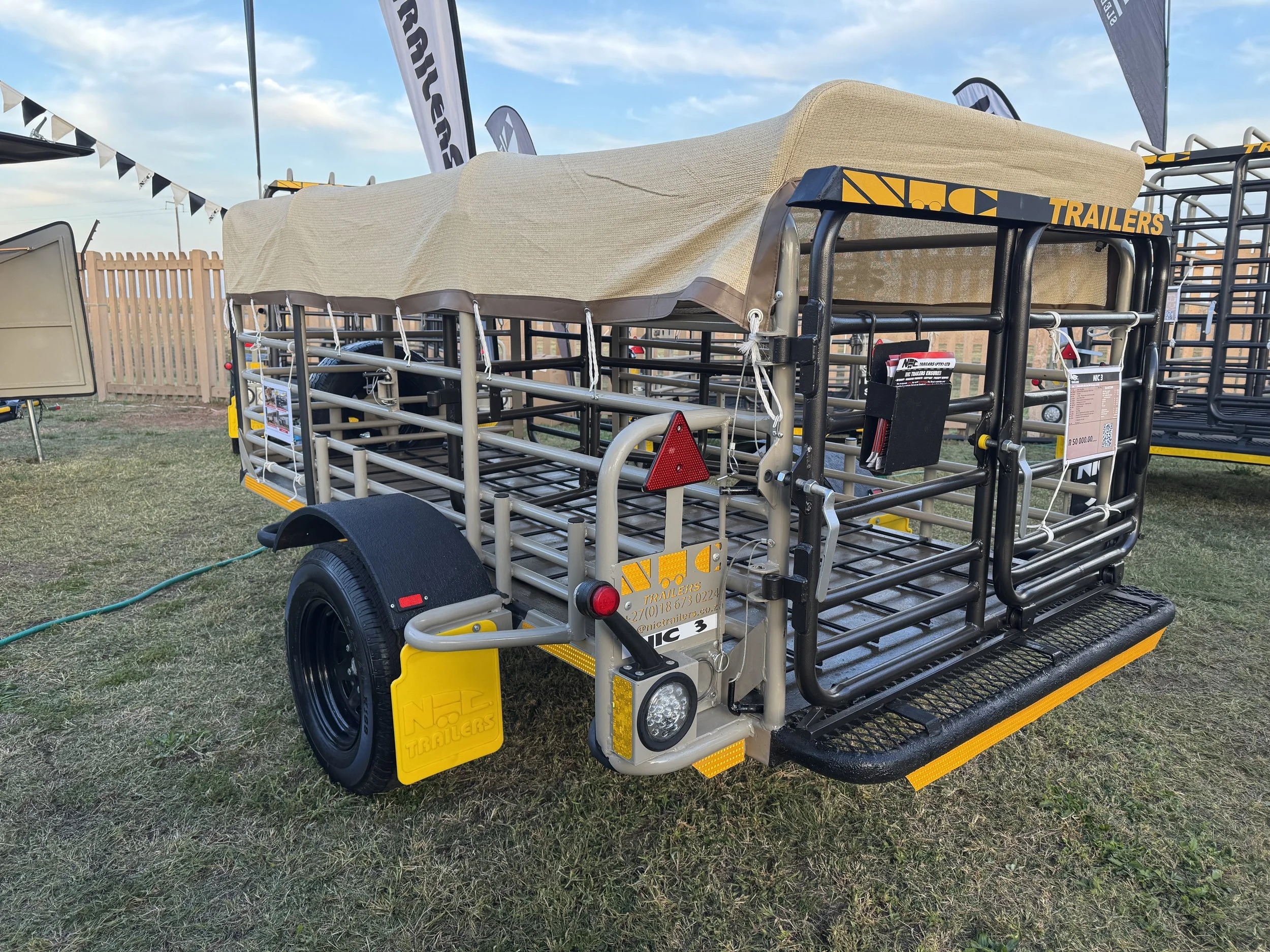 A livestock trailer with a beige canopy on top, parked on grass, surrounded by similar trailers and flags at an outdoor event.