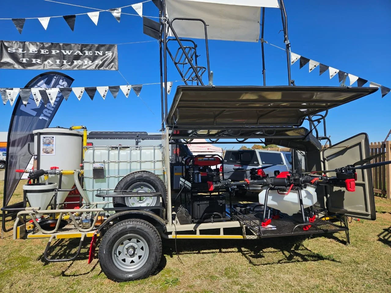 A modified vehicle equipped with outdoor gear and equipment for drone operation and water spraying, parked on grass with a blue sky background.