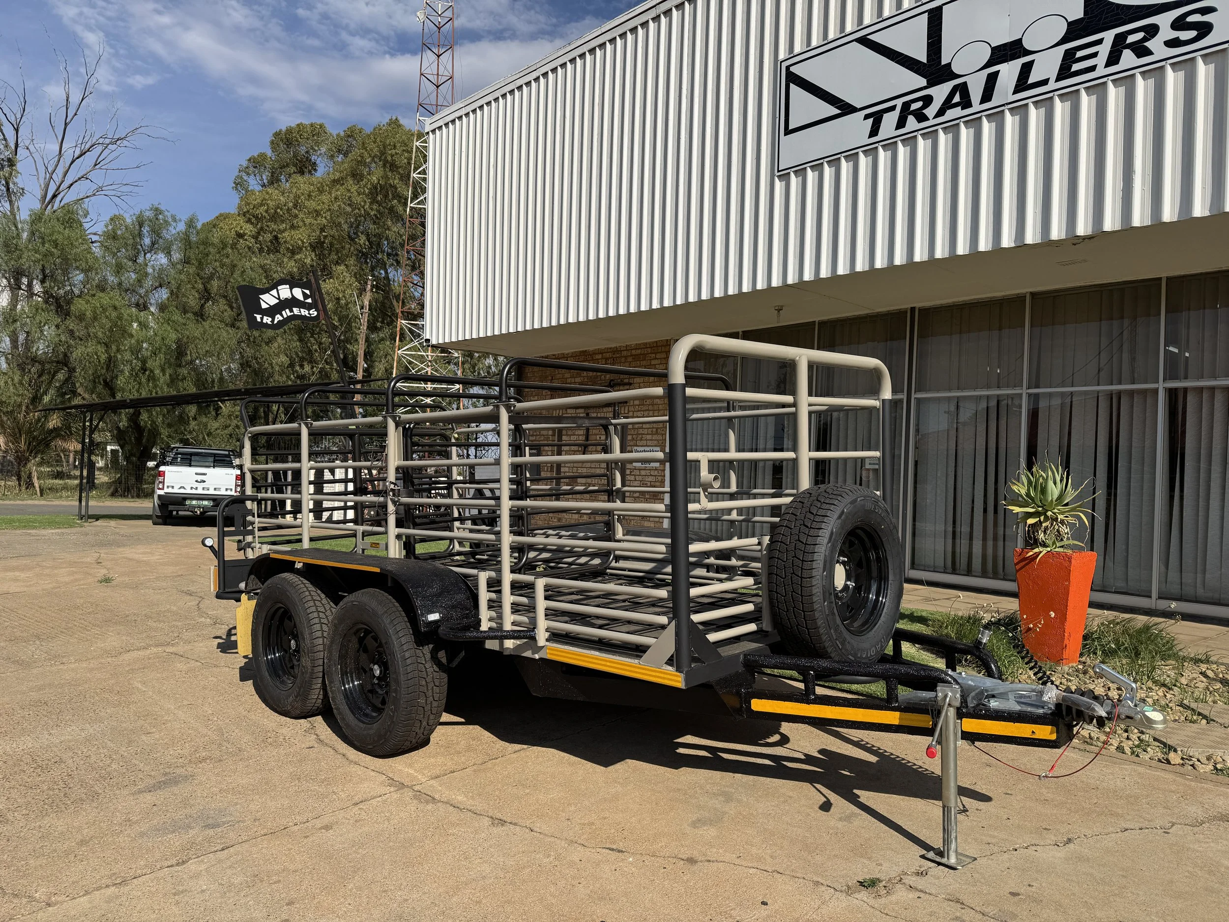 A livestock trailer is parked outside a building with a "Trailers" sign. The trailer has metal railings and spare tires. A potted plant is nearby, and a vehicle is visible in the background.