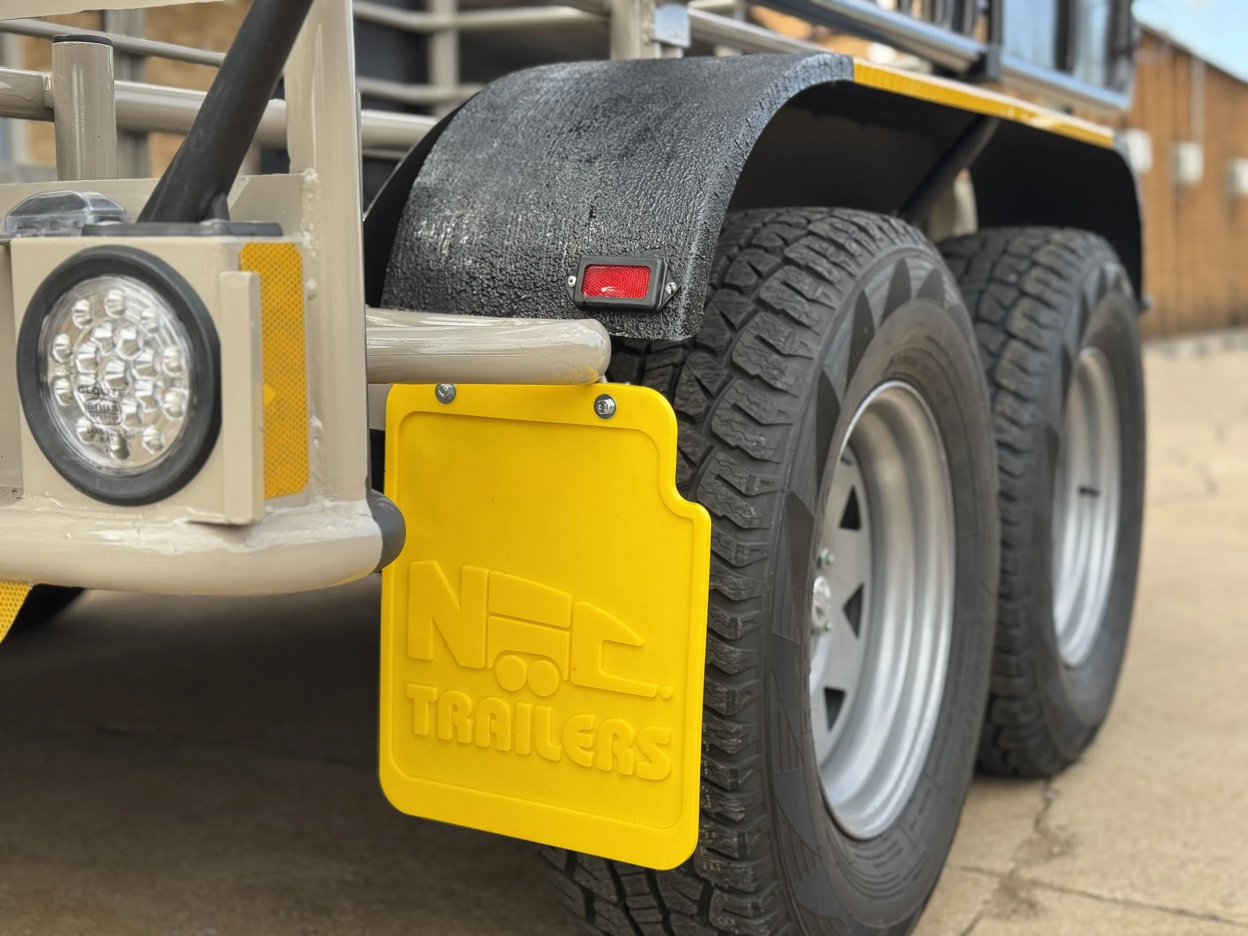 Close-up of a trailer with yellow mud flaps and dual tires. The mud flap has "N Trailers" embossed on it, and there's a round light attached to the trailer's frame.