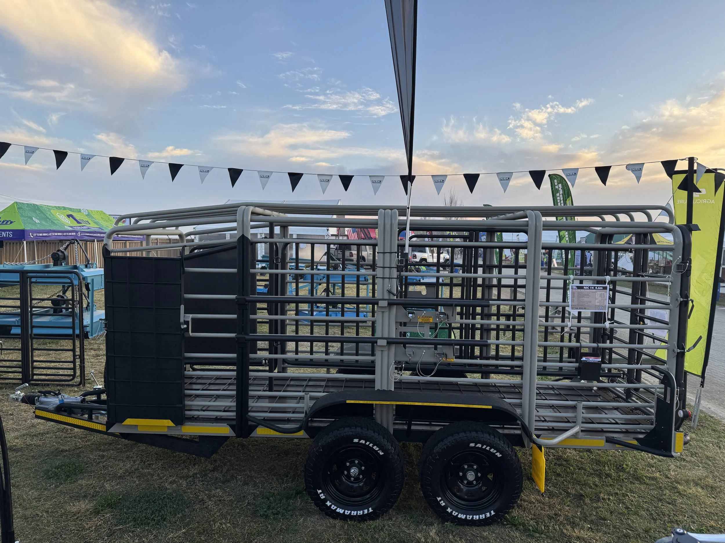 A metal livestock trailer with two axles and black tires parked on grass at an outdoor event. The trailer has a grated floor and side panels and is surrounded by tents and flags, with a partly cloudy sky in the background.