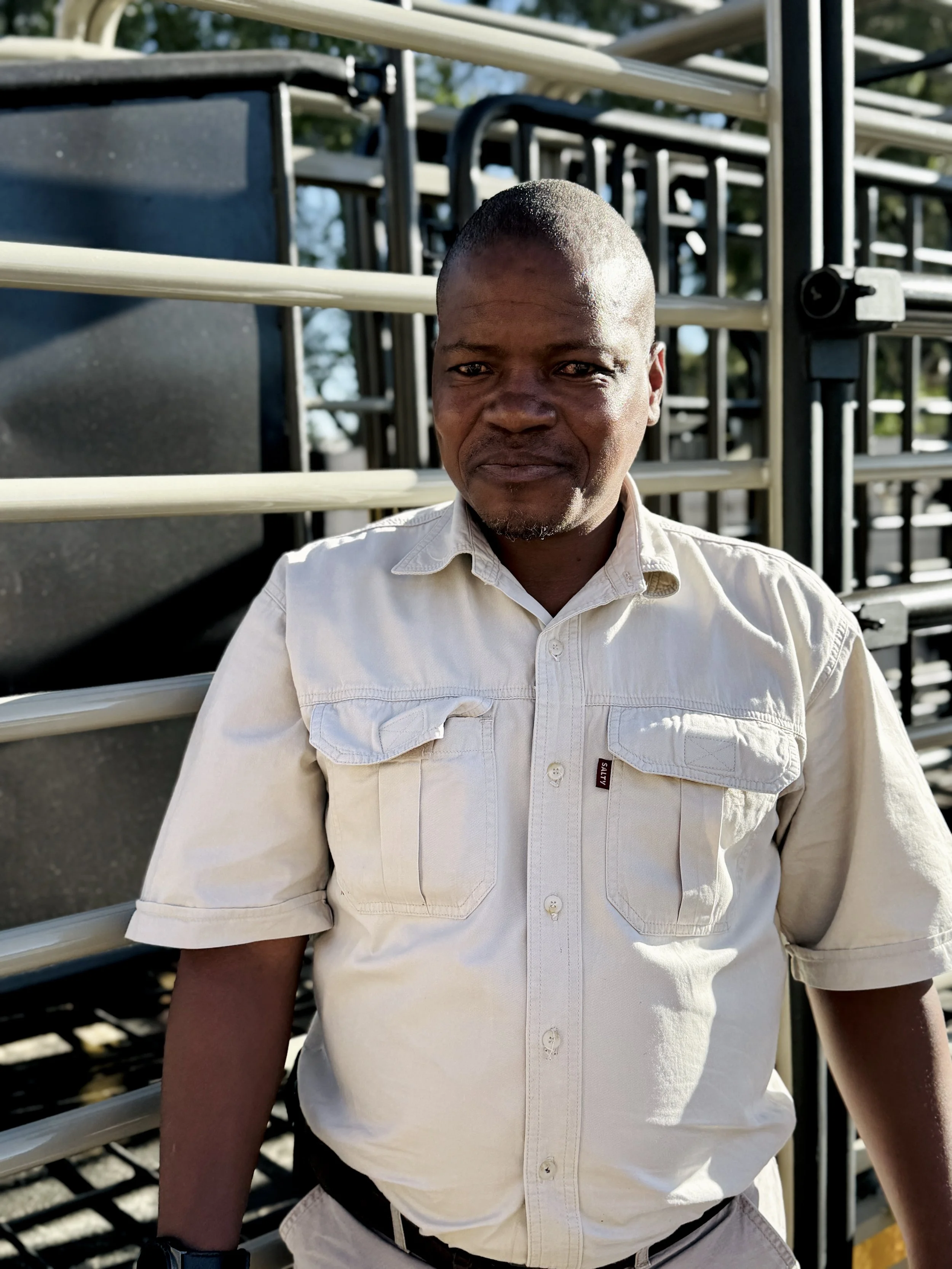 A man with a short haircut and dark skin, wearing a light-colored button-up shirt with chest pockets, standing outdoors in front of stacked black and beige metal chairs or carts, with sunlight illuminating his face.