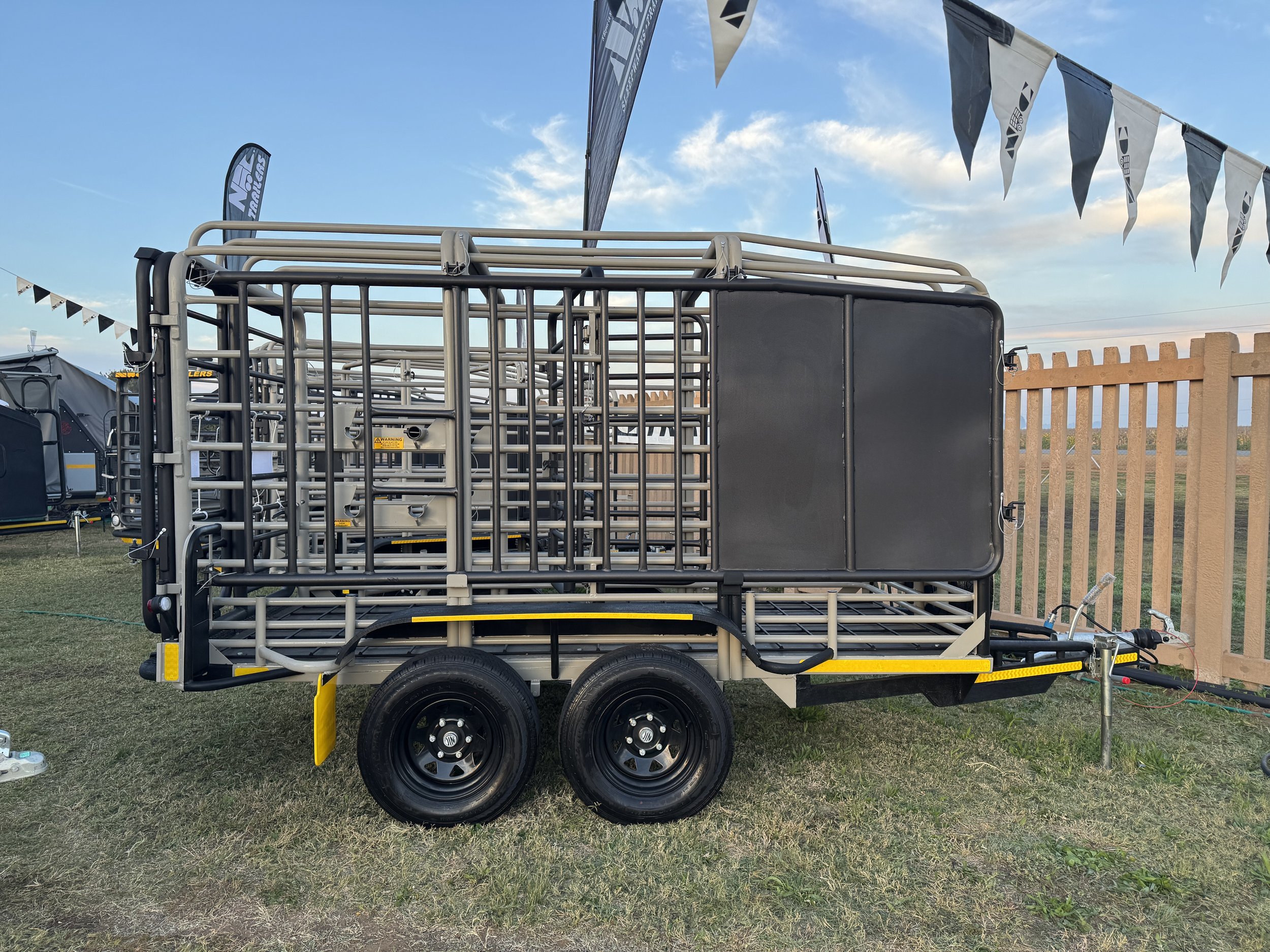 Metal livestock trailer with dual axles parked on grass at an outdoor event, with flags and a wooden fence visible in the background.
