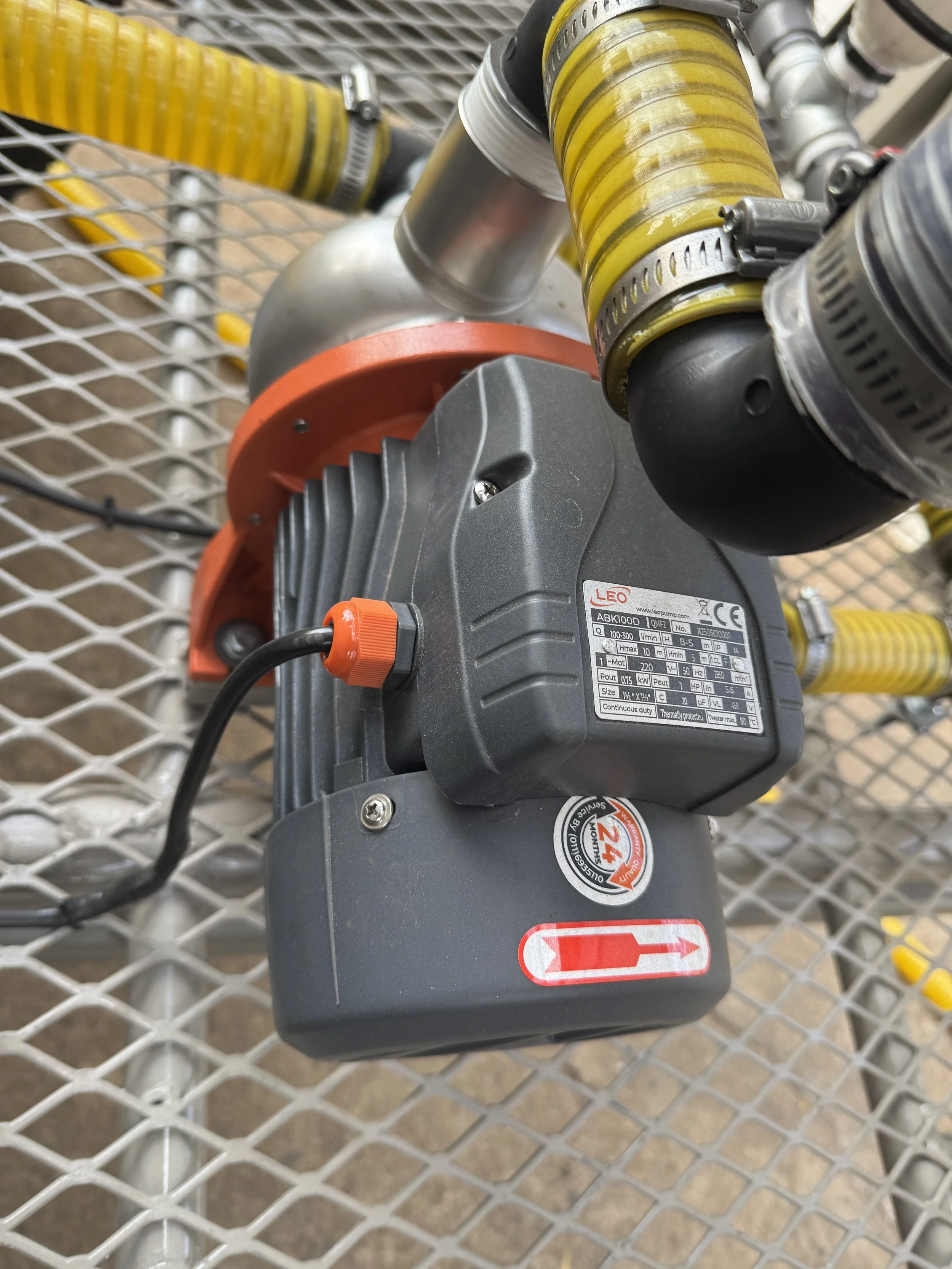 Close-up of industrial equipment with pipes, hoses, and a motor labeled LEO, mounted on a metal grating surface.