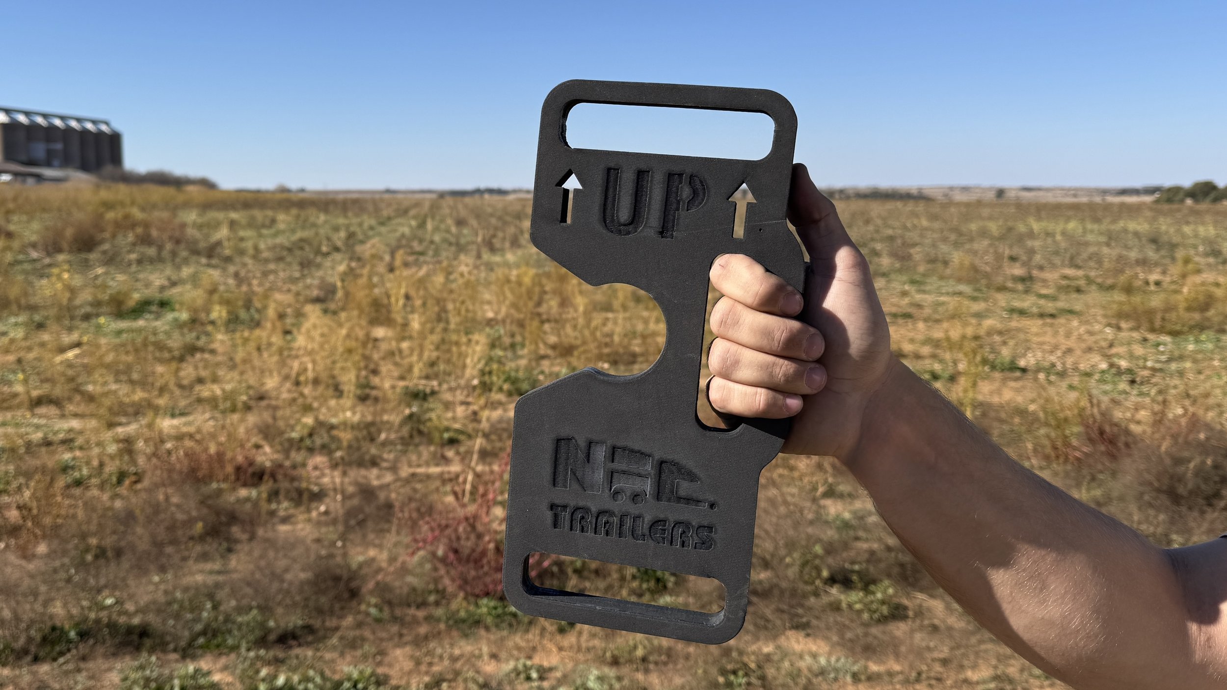 Person holding a black metal sign with cutouts that says 'UP' with arrows and 'N.CE. TRAILERS' in a field of yellow-green grass with a building in the distance under a clear blue sky.