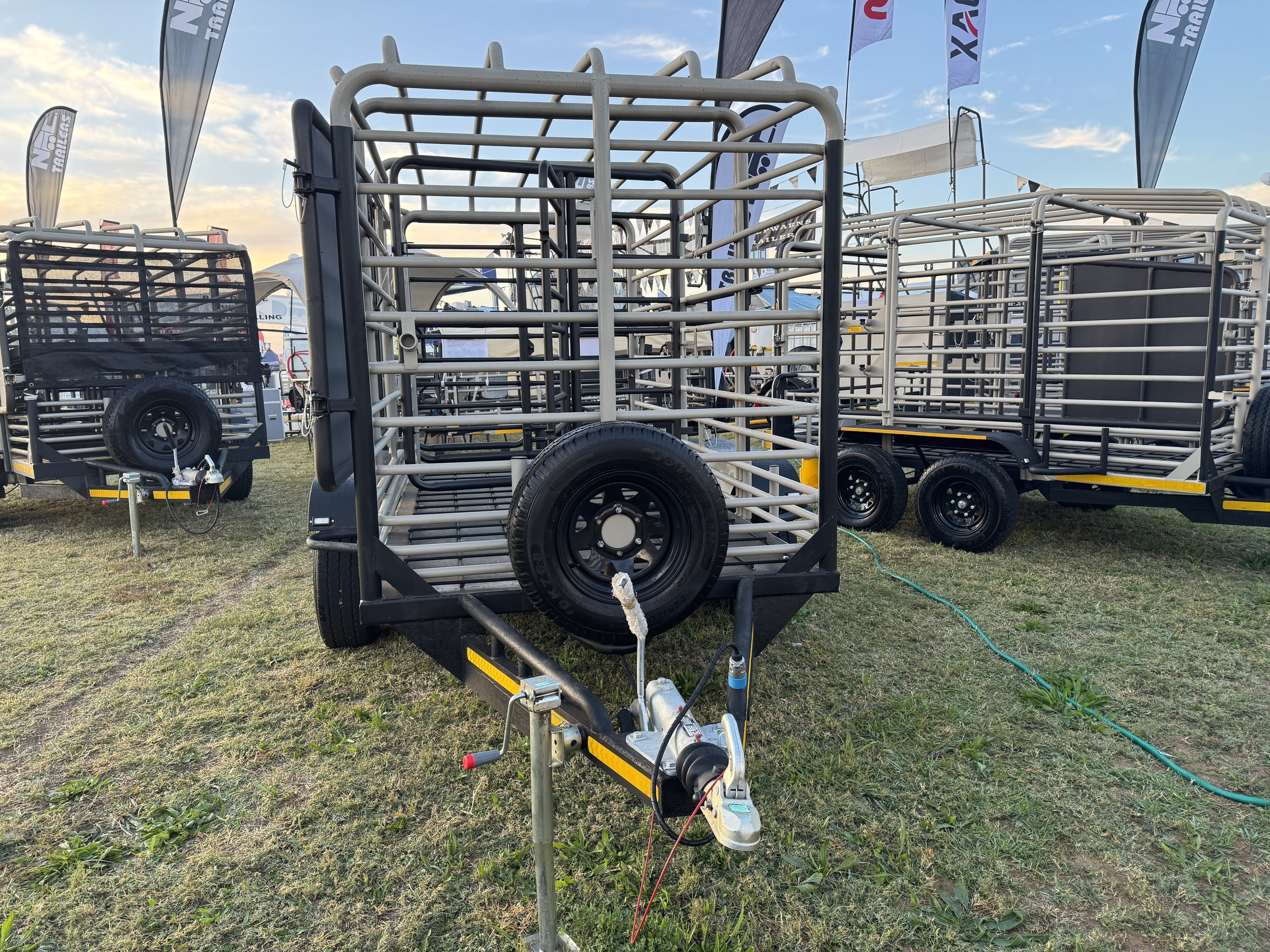 A trailer with metal bars, a spare tire, and a hitch, set on a grassy field with other trailers and flags in the background at an outdoor event.