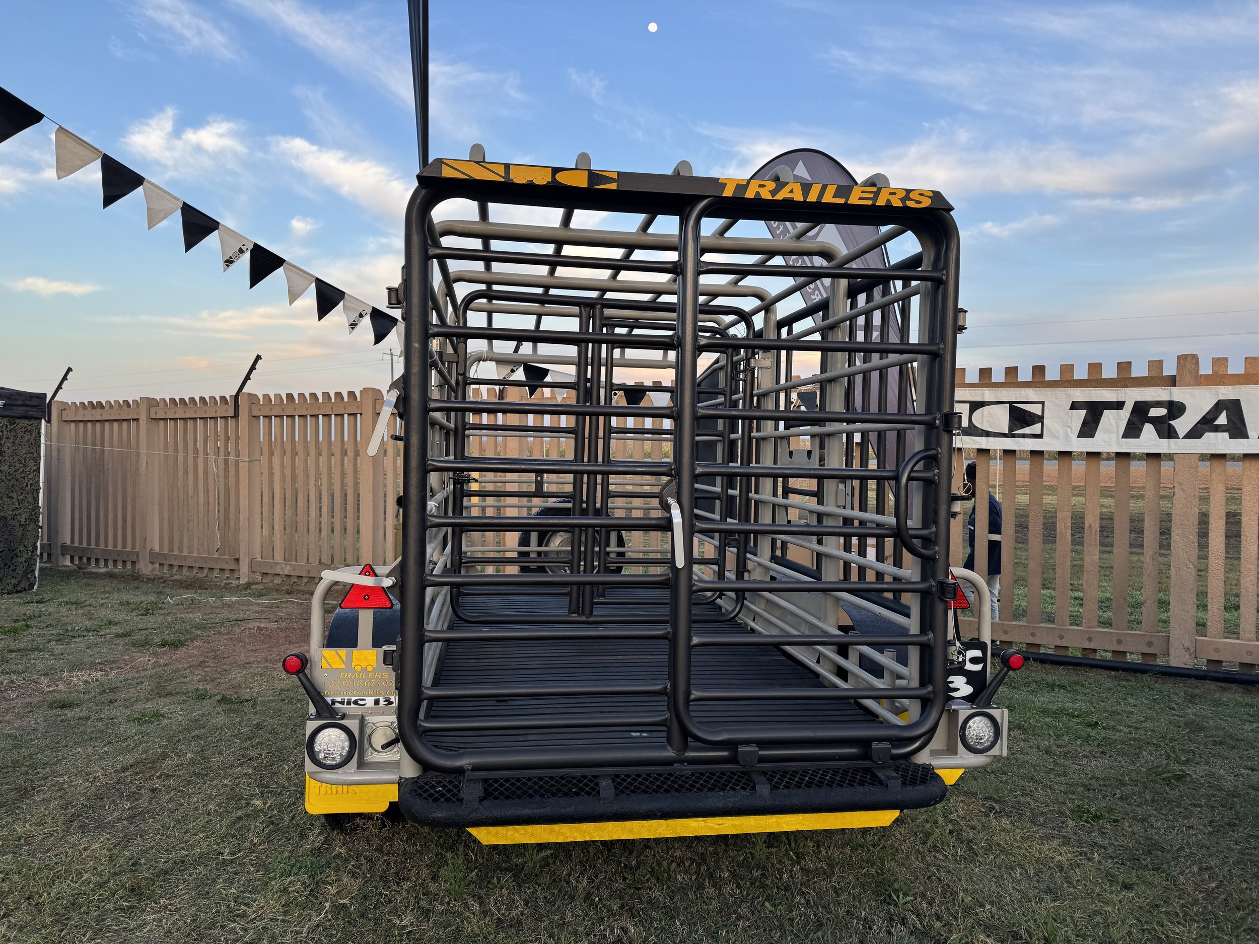 Front view of a flatbed trailer with metal guardrails, parked on grass with a wooden fence and blue sky in the background.
