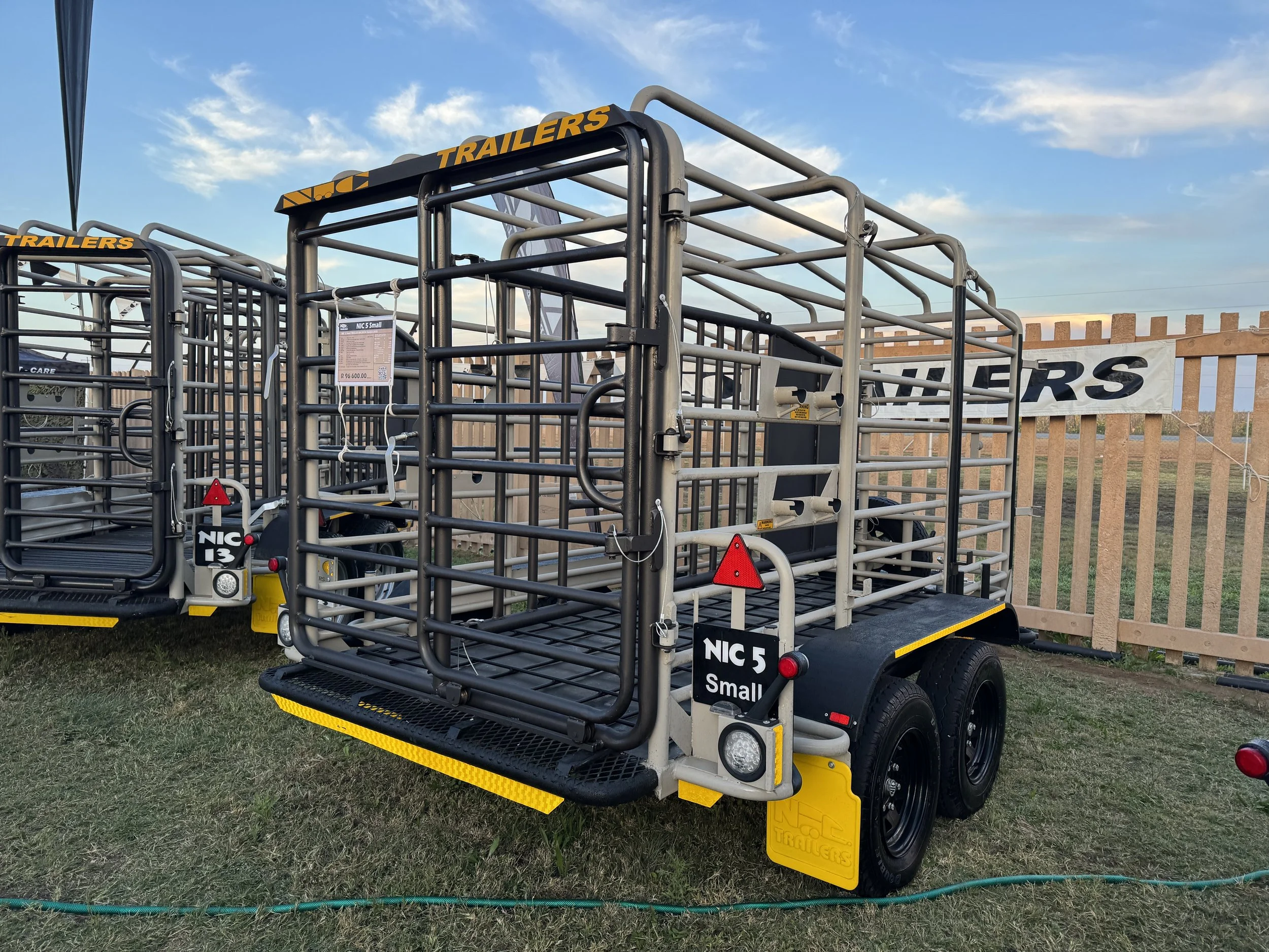 A small, gray utility trailer with metal fencing and a black rubber floor, parked on grass at an outdoor exhibition. The trailer has dual black wheels, a warning light, and a sign that reads 'NIC 5 Small'.