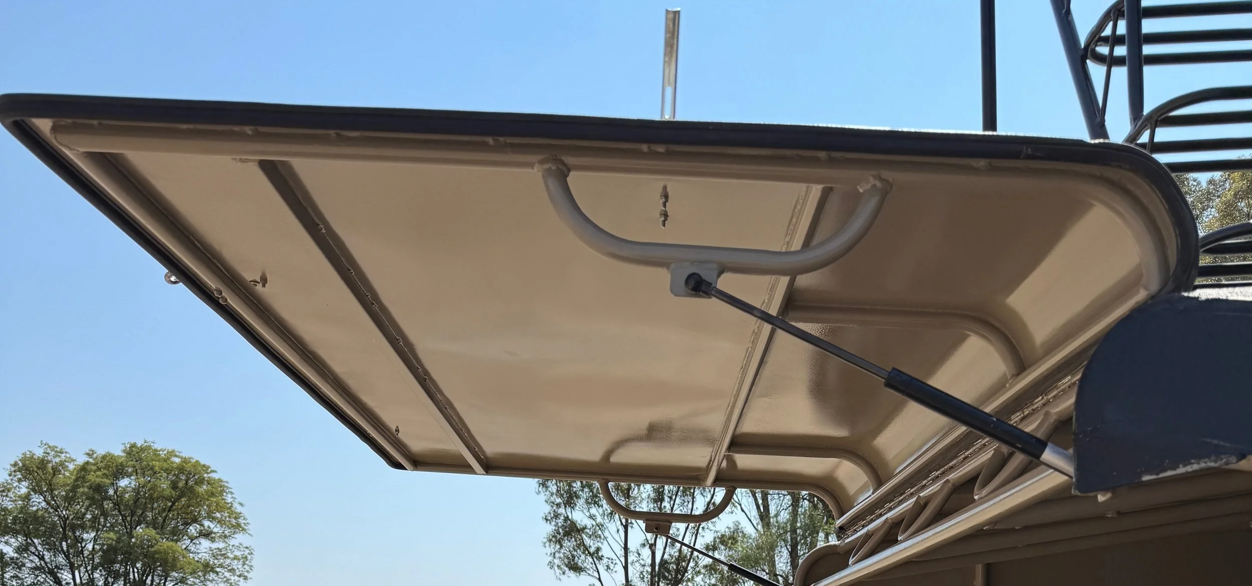 Open hatchback of a vehicle with a hydraulic lift, viewed from below, showing metal frame and latch against a clear blue sky.
