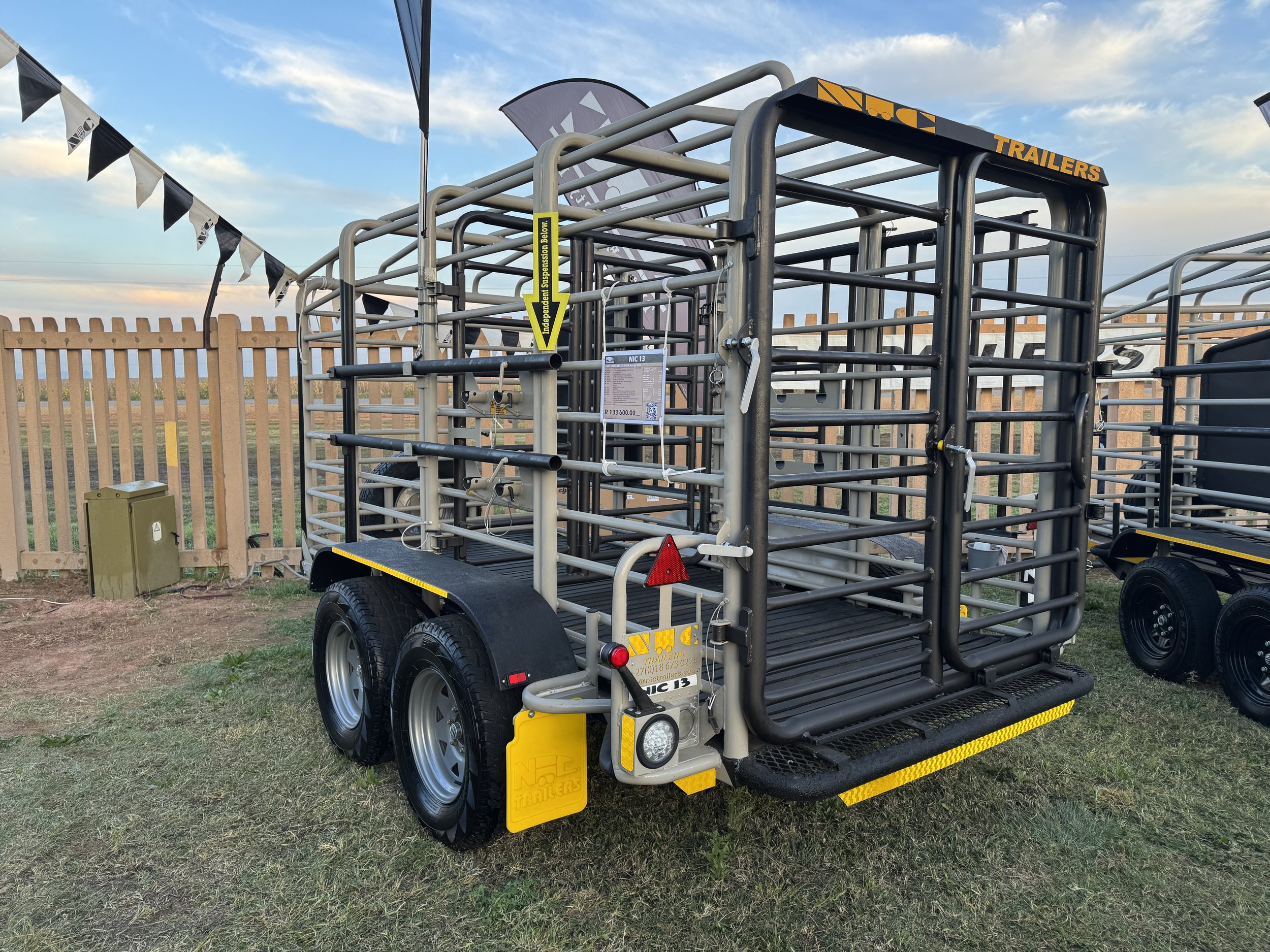 A livestock trailer with a metal frame, two axles, and black rubber tires, parked outdoors on grass with a wooden fence and a partly cloudy sky in the background.
