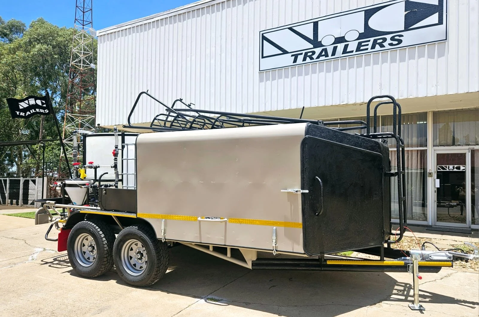 Silver trailer with black framing and dual tires parked outside a building with a sign reading 'NCS Trailers'