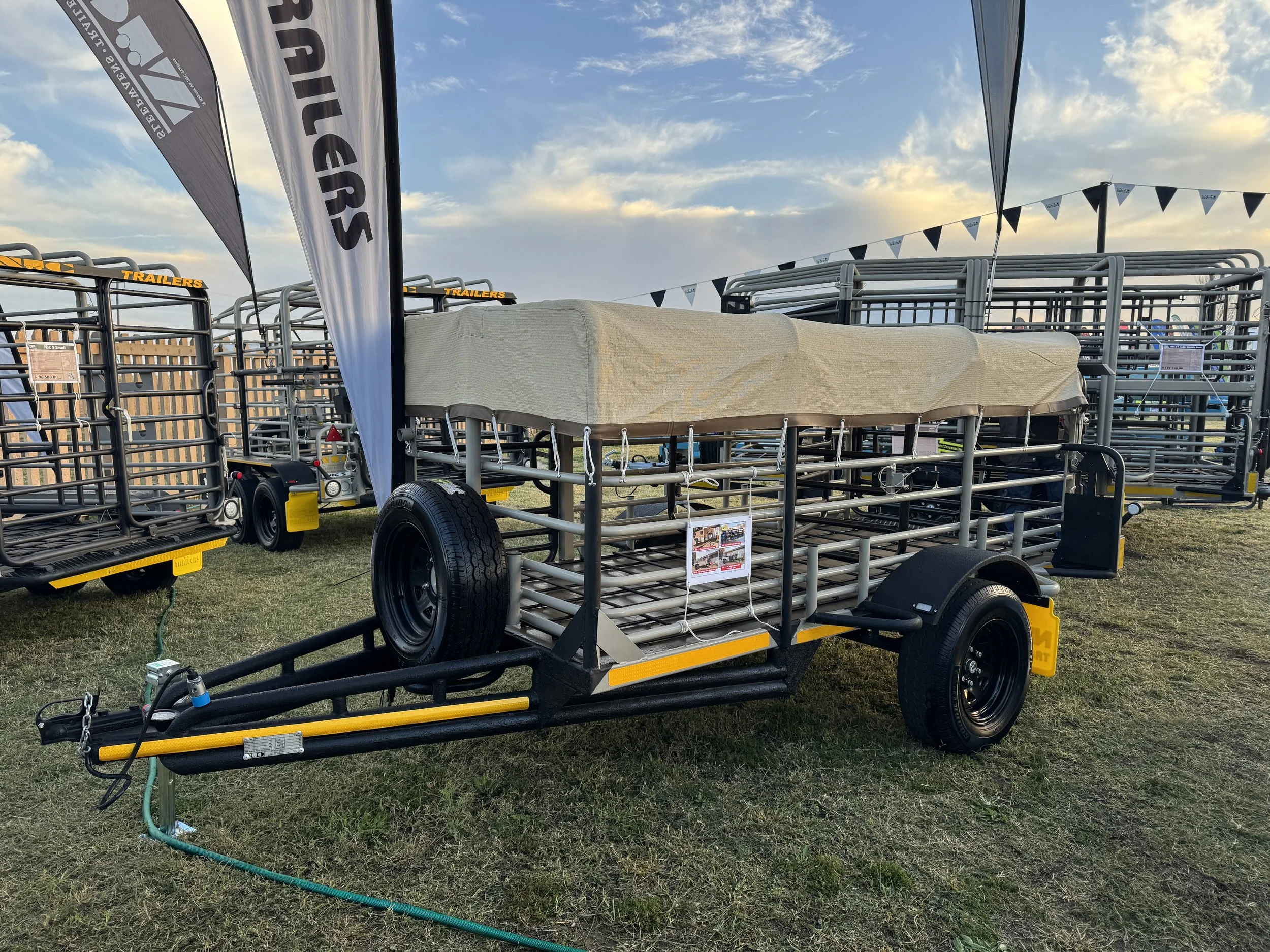 A trailer with a metal frame and large black tires parked on grass at an outdoor equipment sale, with other trailers in the background under a partly cloudy sky.